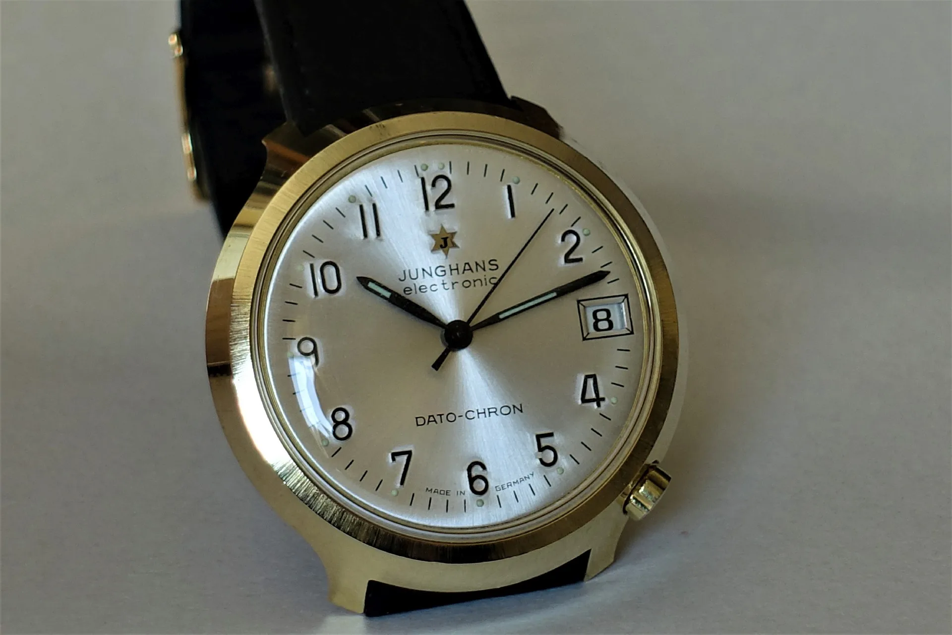 Junghans Dato-Chron Uhr Analoge Armbanduhr mit hellem Zifferblatt, schwarzem Lederarmband und goldfarbenem Gehäuse.
