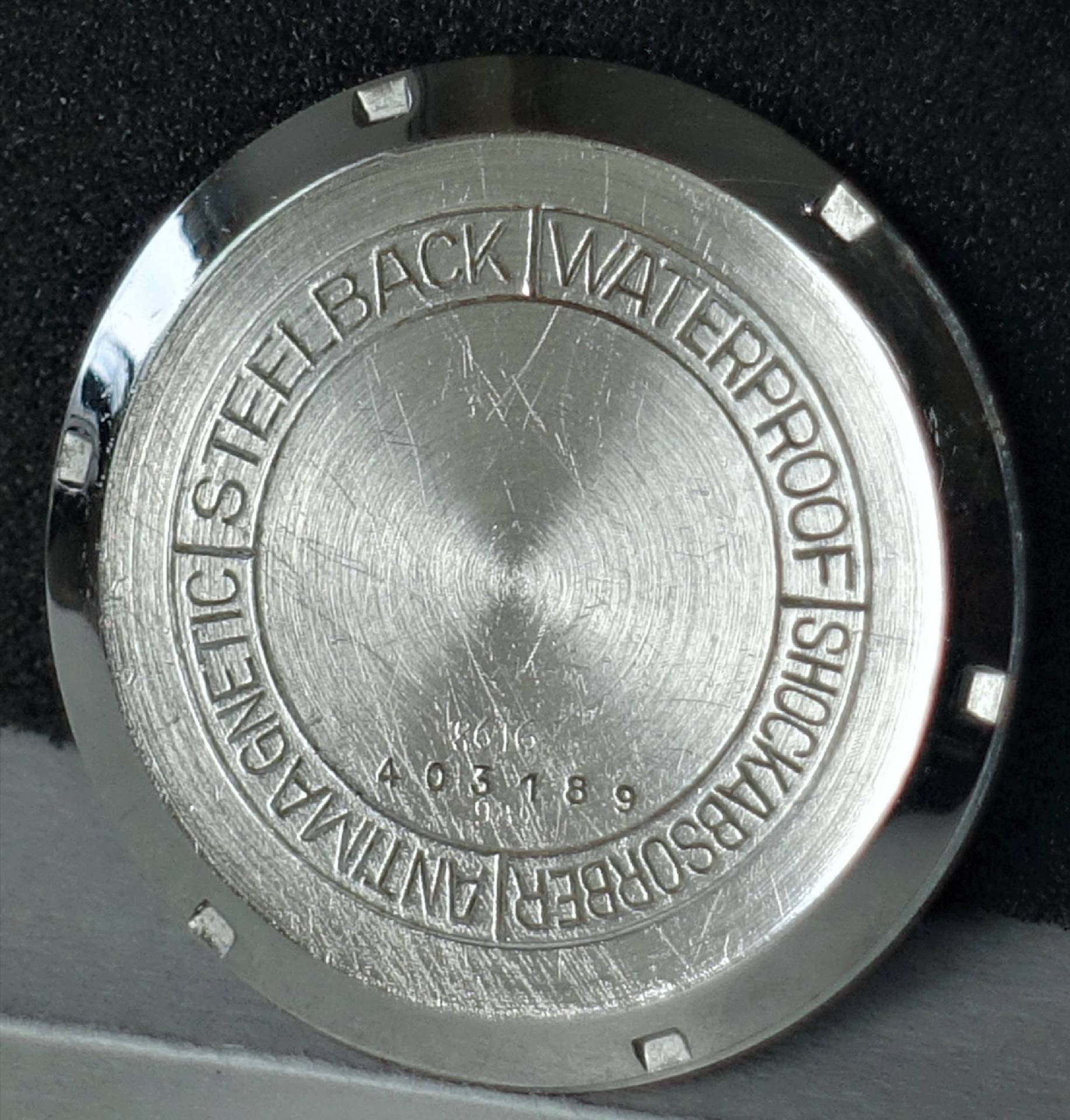 Runder silberfarbener Uhrenboden mit Gravur und Kerben am Rand.