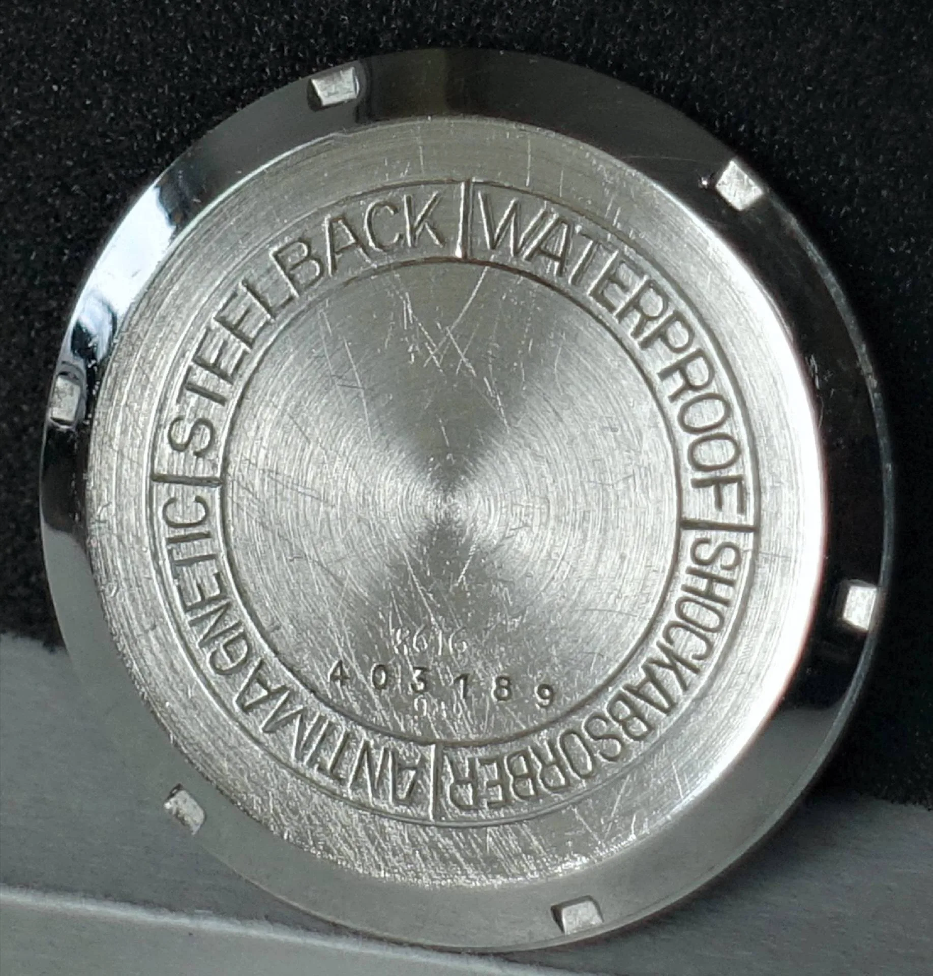 Runder silberfarbener Uhrenboden mit Gravur und Kerben am Rand.