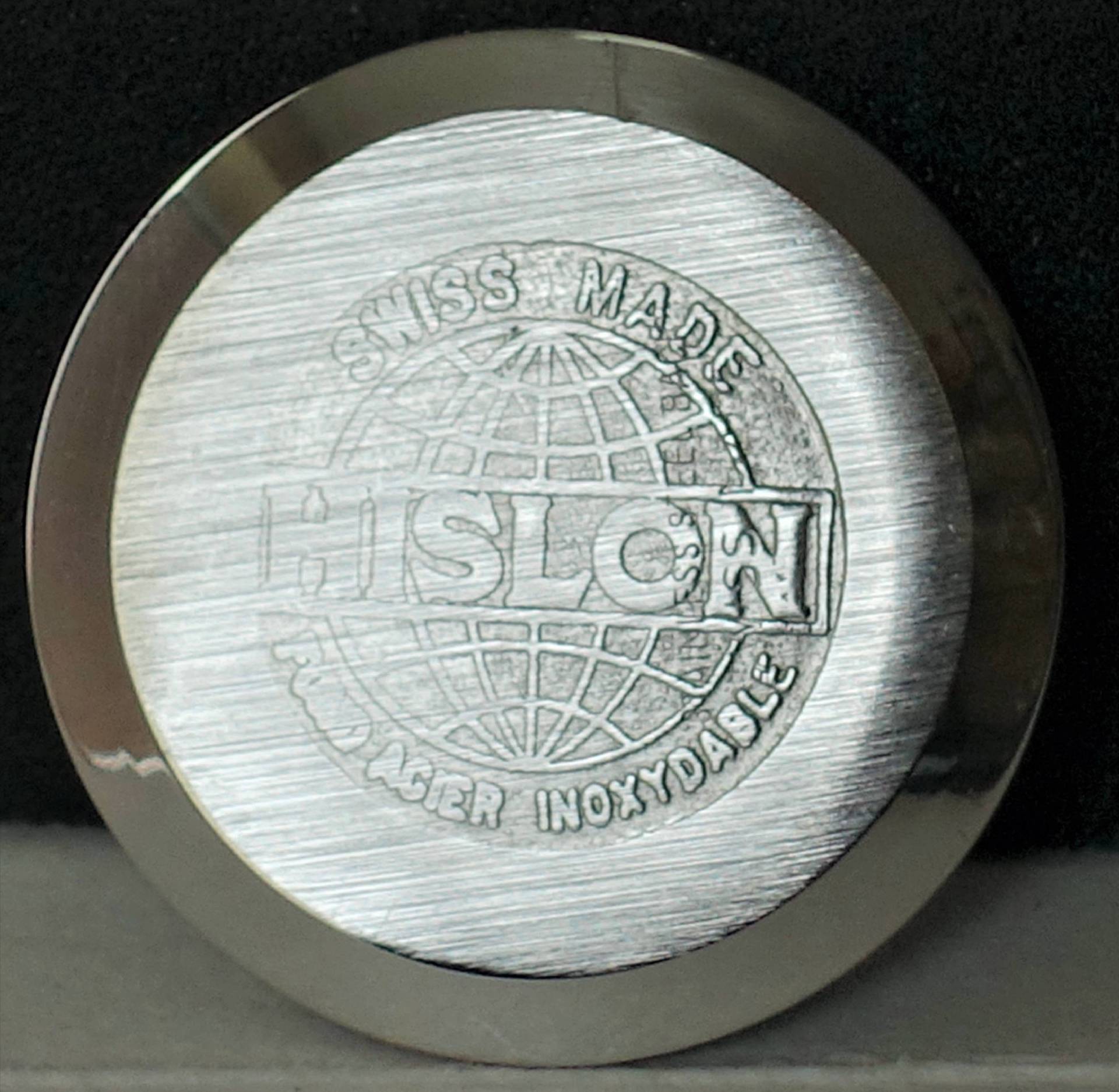 Rundes, silberfarbenes Uhrenboden-Bauteil mit Gravur des Schriftzugs "HISLON", Weltkugel-Motiv und den Aufschriften "SWISS MADE" sowie "FOND ACIER INOXYDABLE".