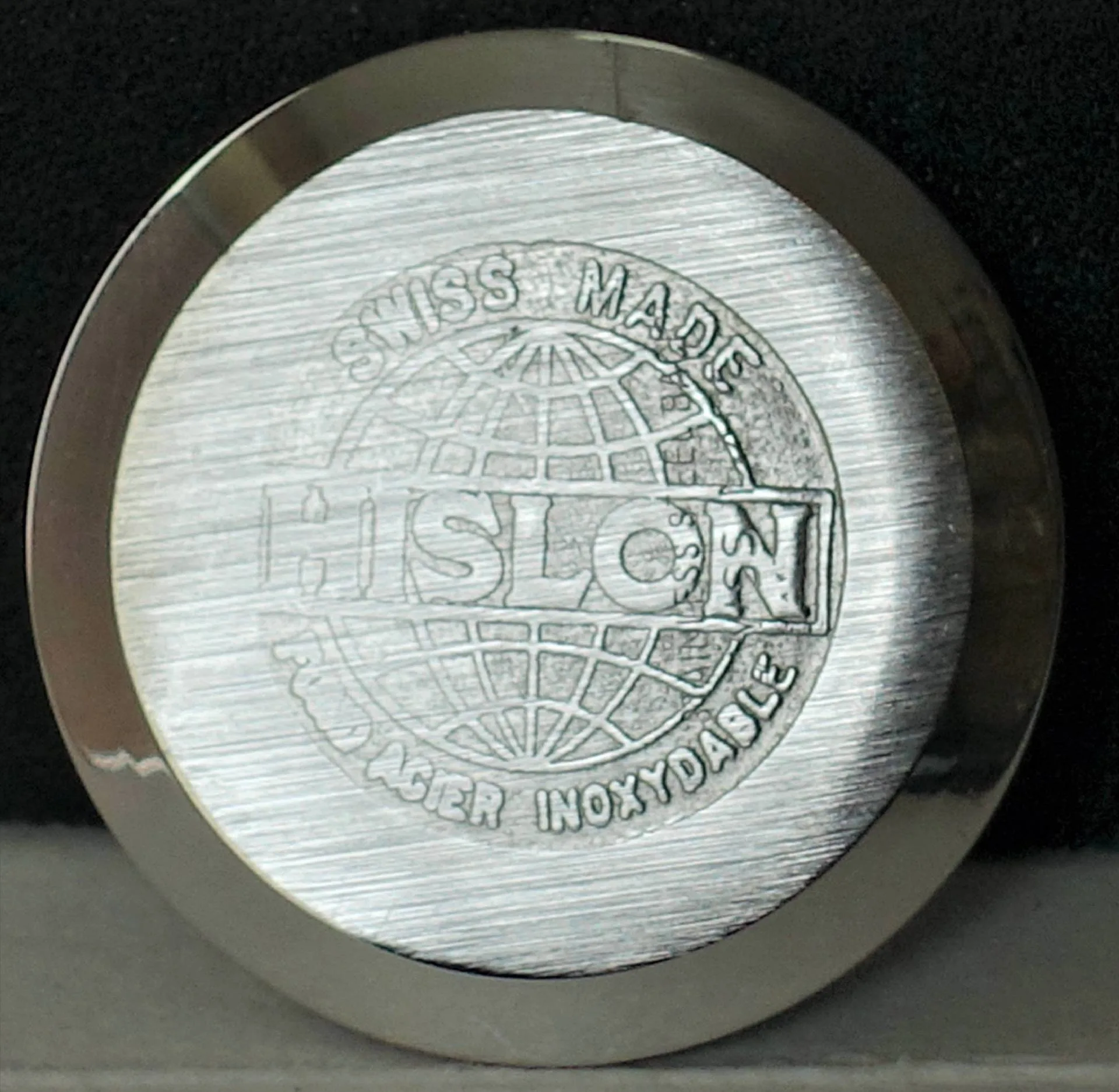 Rundes, silberfarbenes Uhrenboden-Bauteil mit Gravur des Schriftzugs "HISLON", Weltkugel-Motiv und den Aufschriften "SWISS MADE" sowie "FOND ACIER INOXYDABLE".