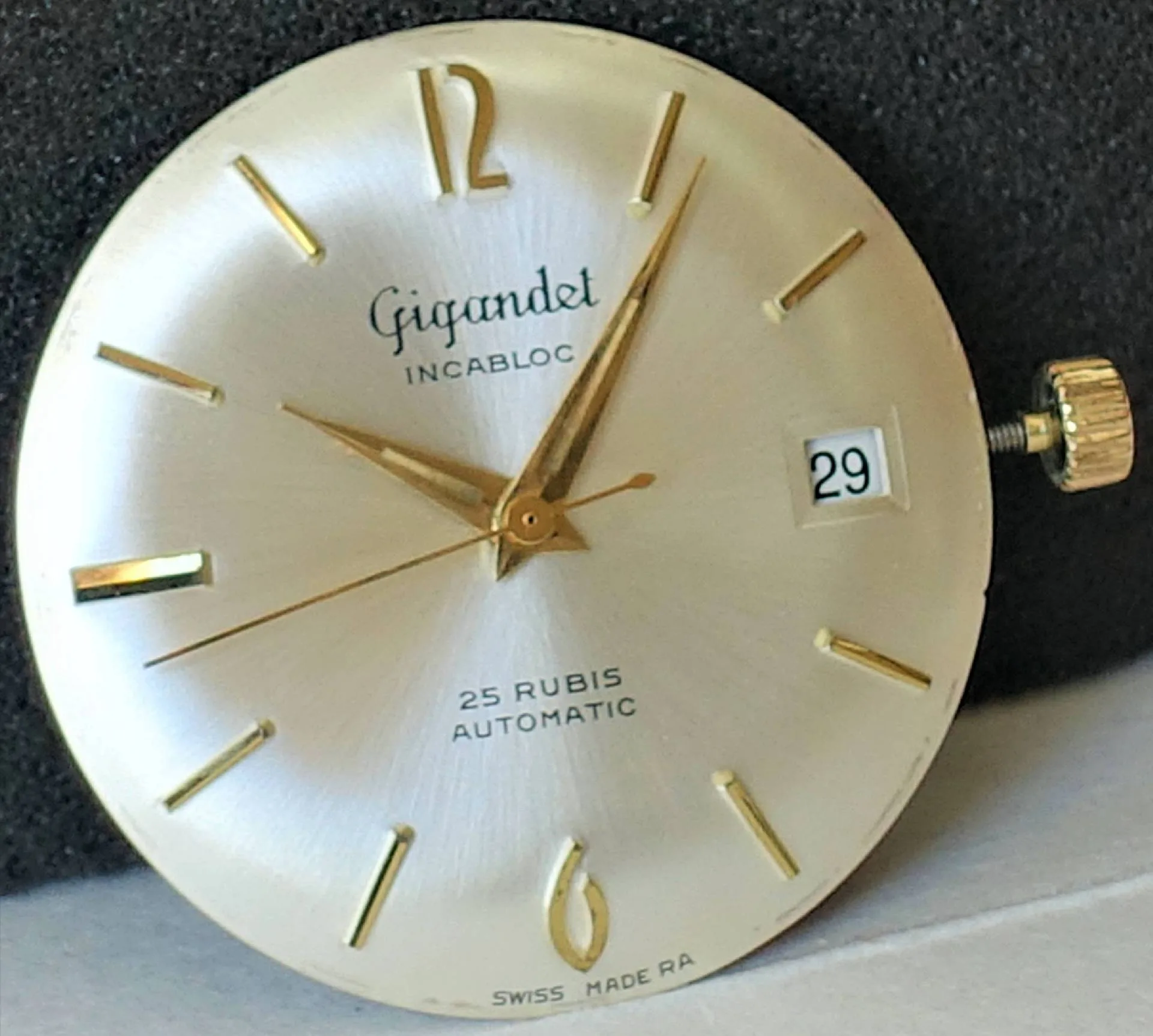 Silbernes Zifferblatt goldene Indizes Rundes silberfarbenes Uhrwerk mit goldfarbenen Zeigern und Indizes, Datumsanzeige bei drei Uhr, Aufschrift „Gigandet Incabloc 25 Rubis Automatic“.
