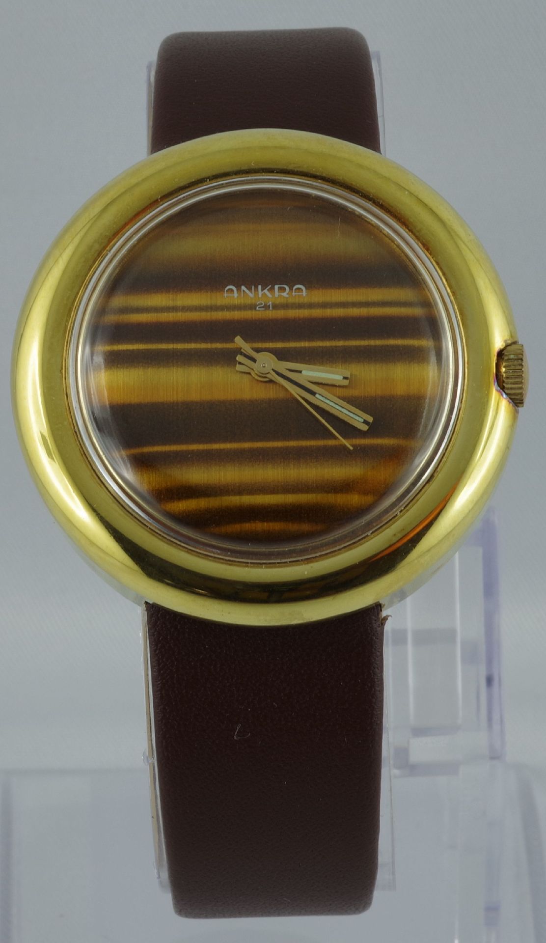 Analoge Armbanduhr mit goldfarbenem Gehäuse, braunem Lederarmband und gestreiftem Zifferblatt.