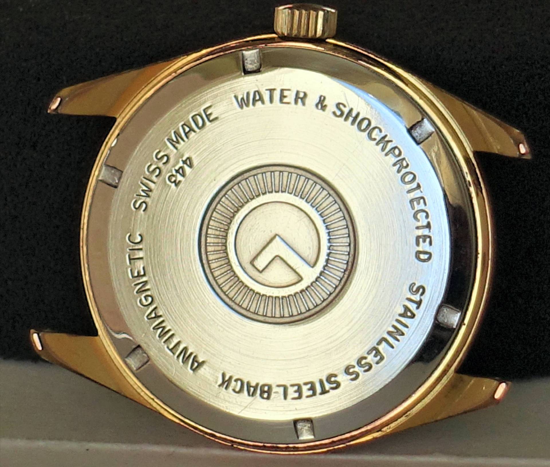 Runder, goldfarbener Gehäuseboden einer Armbanduhr mit silberner Rückseite und eingravierten Beschriftungen.