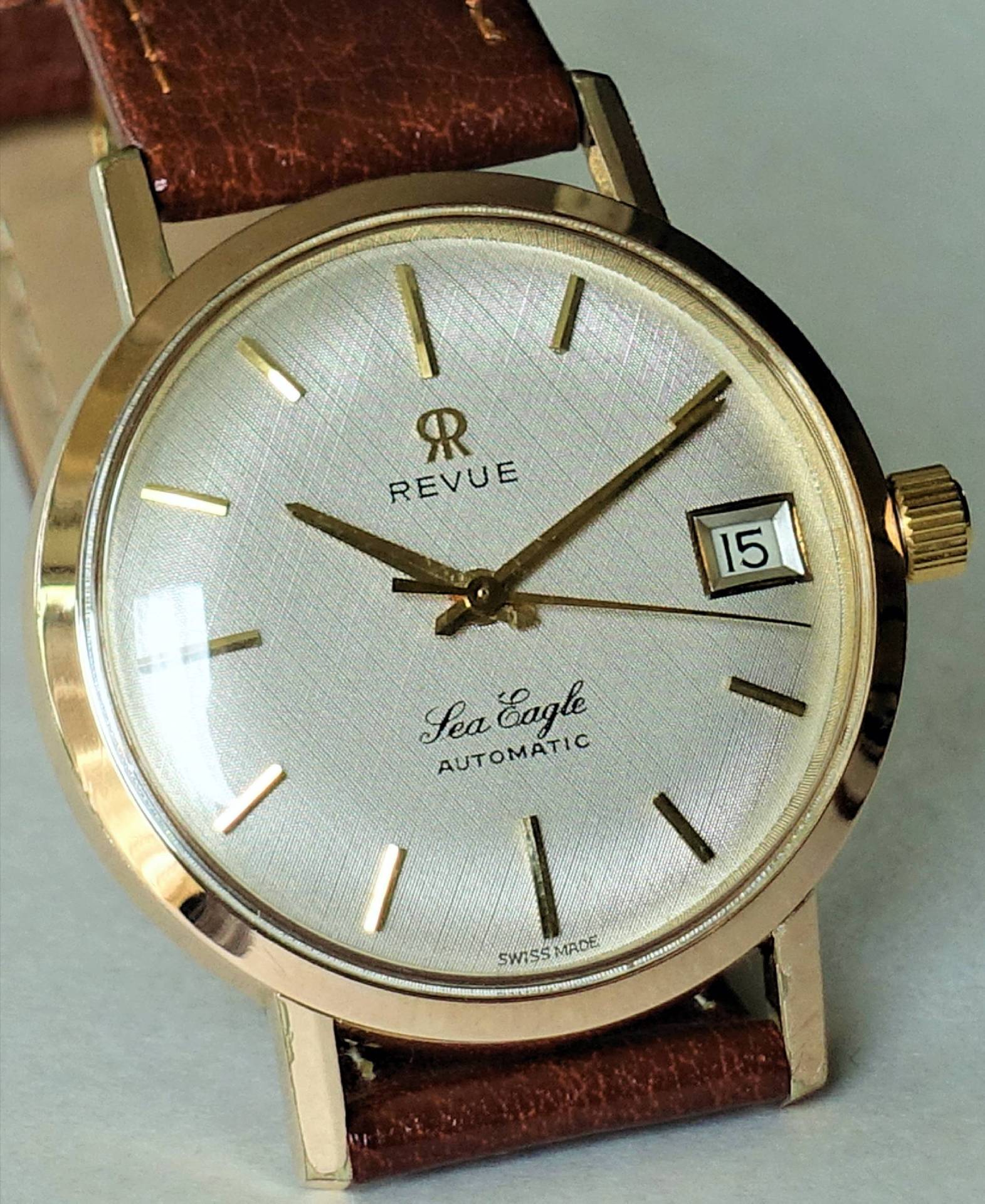 Goldfarbene Armbanduhr mit braunem Armband und silbernem Zifferblatt, Datumsanzeige bei drei Uhr.