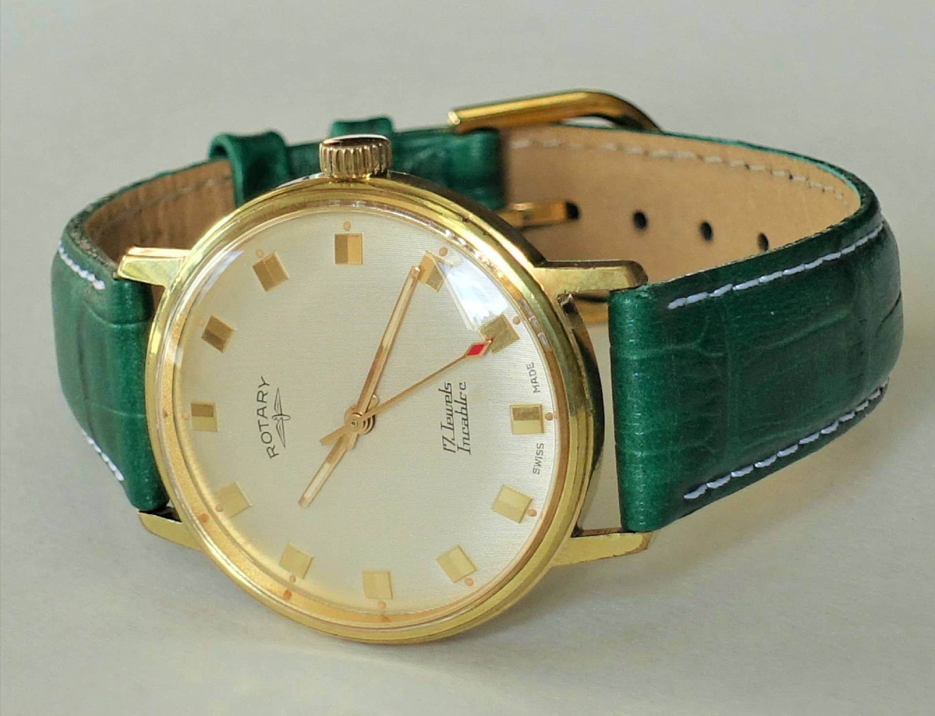 Eine klassische Armbanduhr mit goldfarbenem Gehäuse, hellem Zifferblatt und grünem Lederarmband liegt auf einer hellen Oberfläche.
