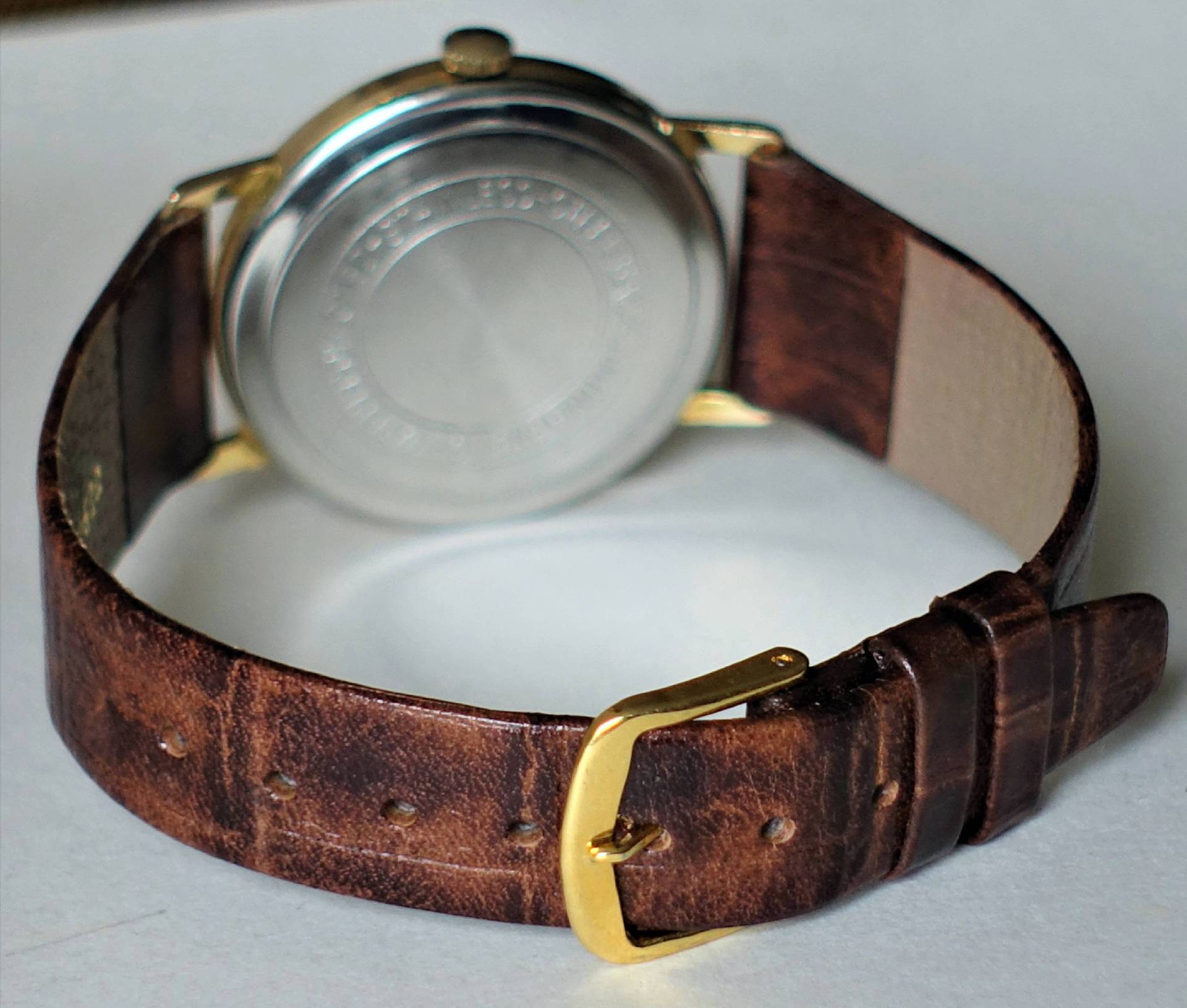 Armbanduhr mit braunem Lederarmband, goldfarbener Schließe und silberfarbener Rückseite.
