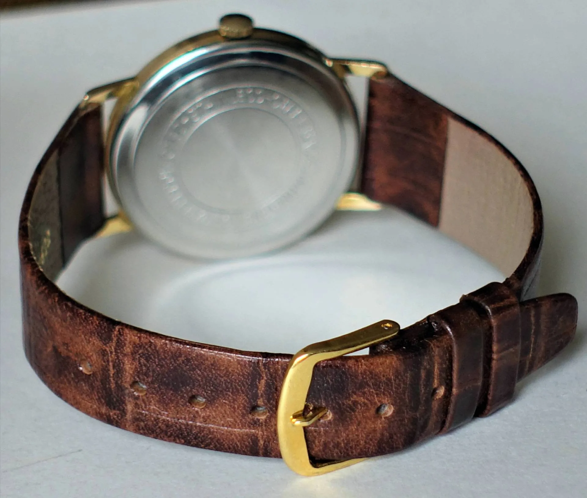 Armbanduhr mit braunem Lederarmband, goldfarbener Schließe und silberfarbener Rückseite.
