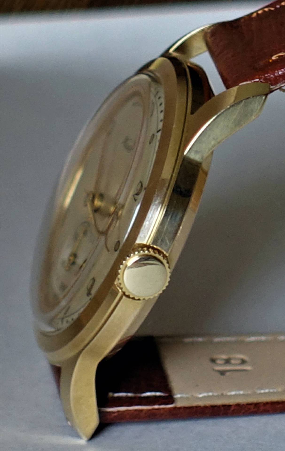 Goldfarbene Armbanduhr mit braunem Lederarmband und seitlicher Krone.