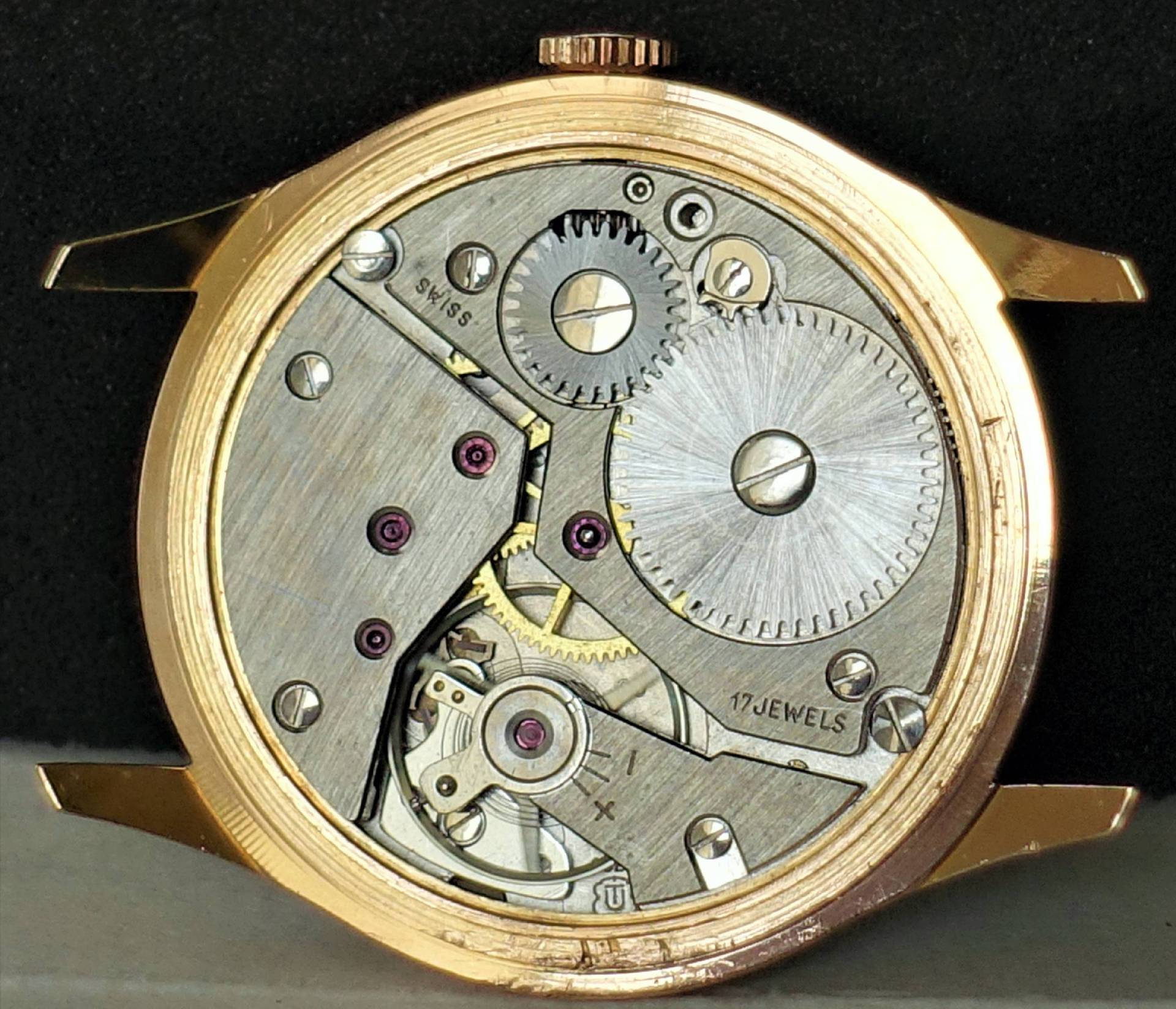 Das Bild zeigt das geöffnete Gehäuse einer Armbanduhr mit einem silberfarbenen mechanischen Uhrwerk und goldfarbenem Gehäuserand.