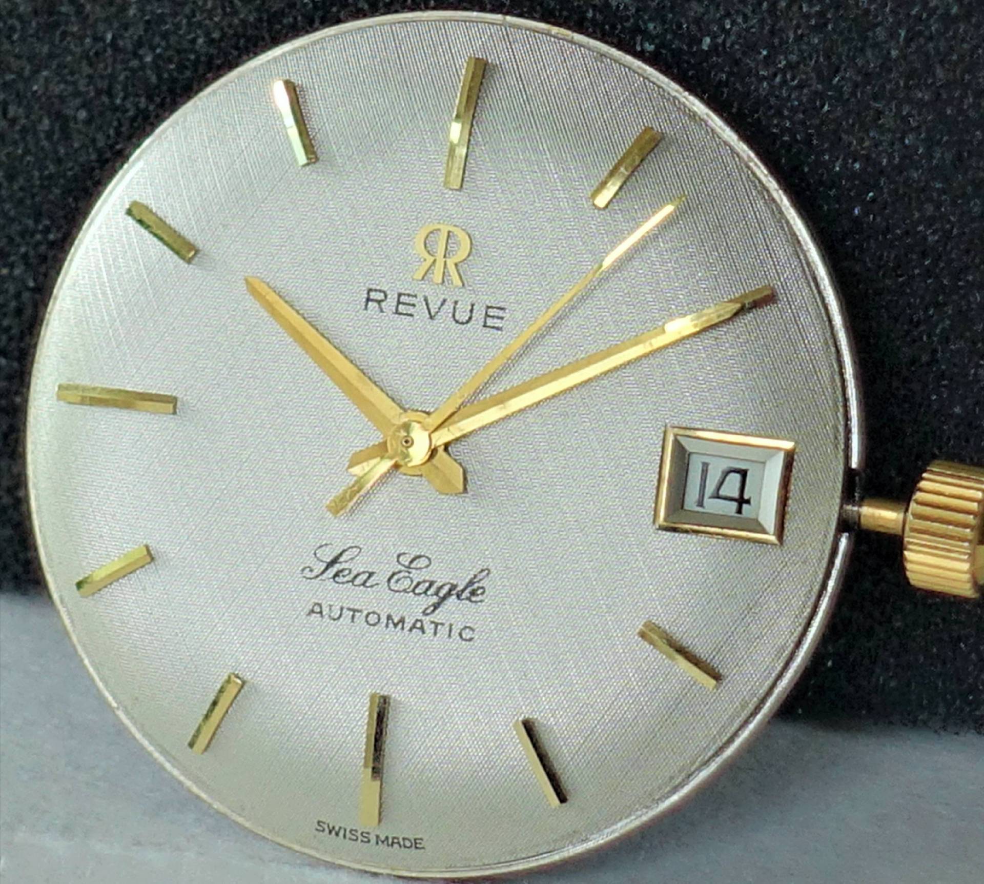 Silberfarbenes Zifferblatt mit goldfarbenen Zeigern und Indizes, Datumsanzeige bei drei Uhr.