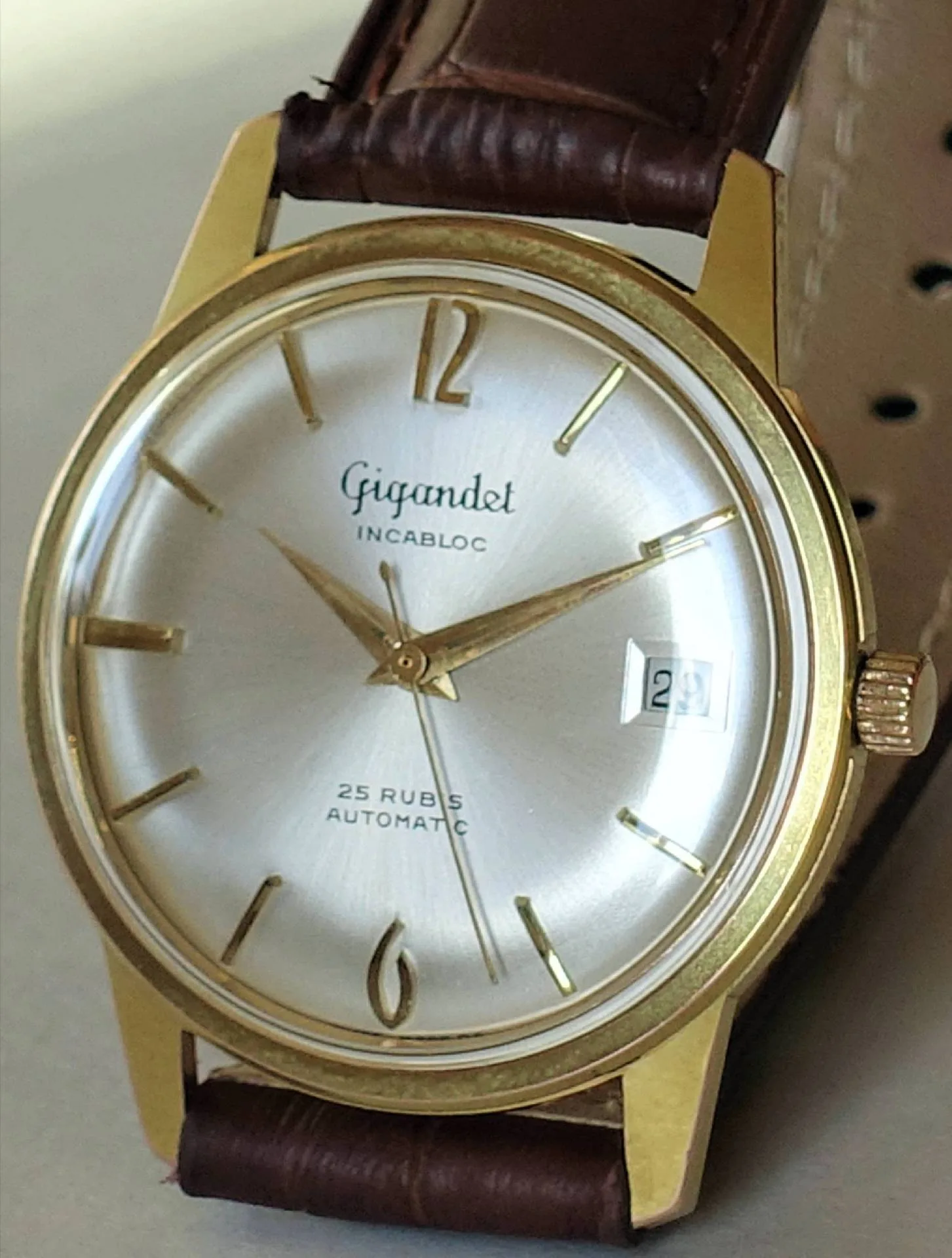 Eine klassische Armbanduhr mit goldfarbenem Gehäuse, silberfarbenem Zifferblatt, braunem Lederarmband und Datumsanzeige bei drei Uhr.