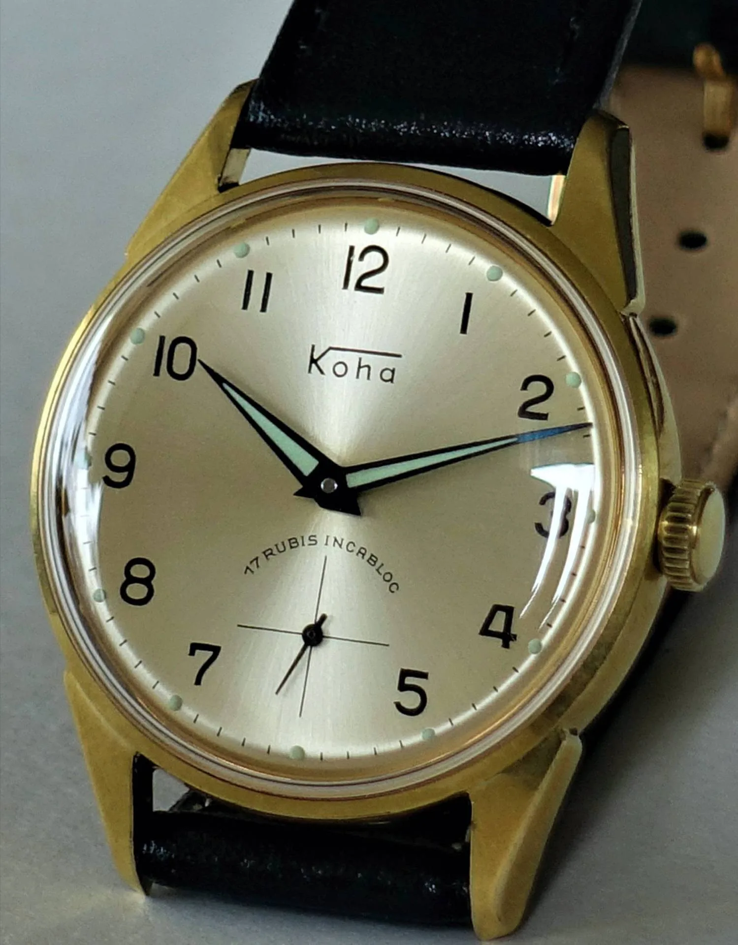 Goldfarbene Armbanduhr mit hellem Zifferblatt, schwarzen Zahlen und schwarzem Lederarmband.