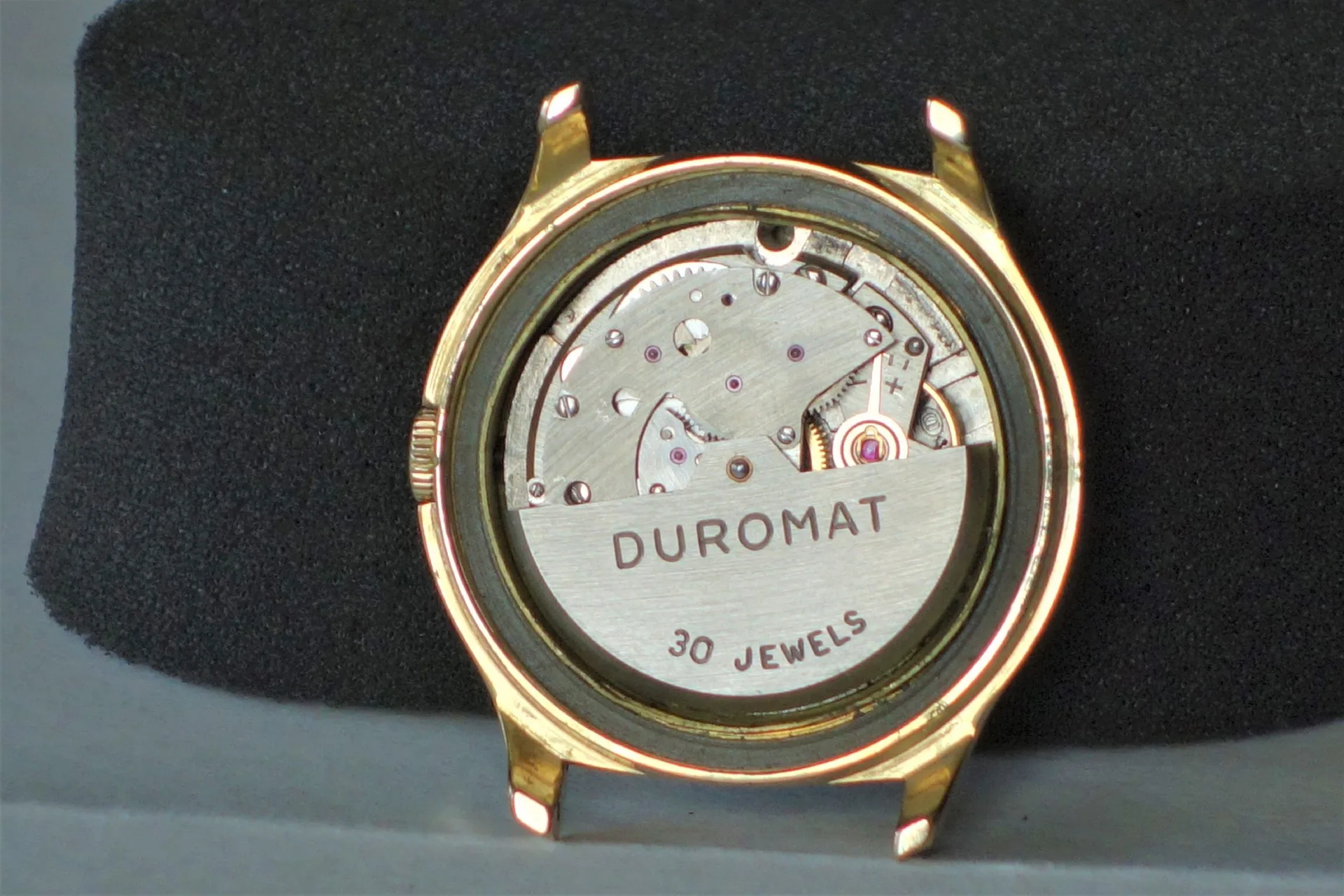 Uhrwerk Duromat 30 Jewels Silberfarbenes Uhrwerk mit der Aufschrift "DUROMAT 30 JEWELS" in einem goldfarbenen Uhrengehäuse.