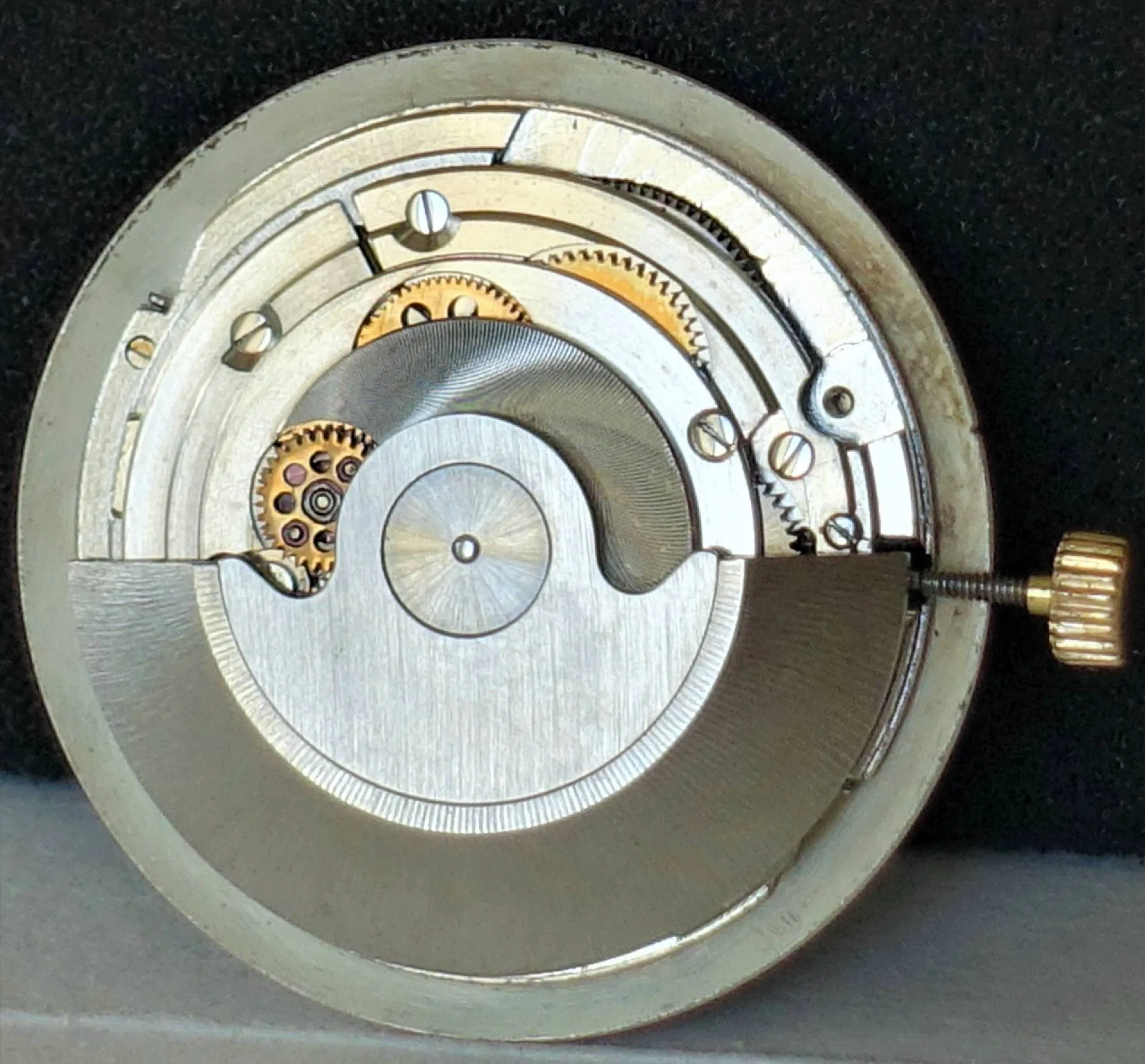 Ein rundes Uhrwerk mit silberfarbenen und goldfarbenen Metallteilen sowie einer goldfarbenen Aufzugskrone ist vor schwarzem Hintergrund zu sehen.