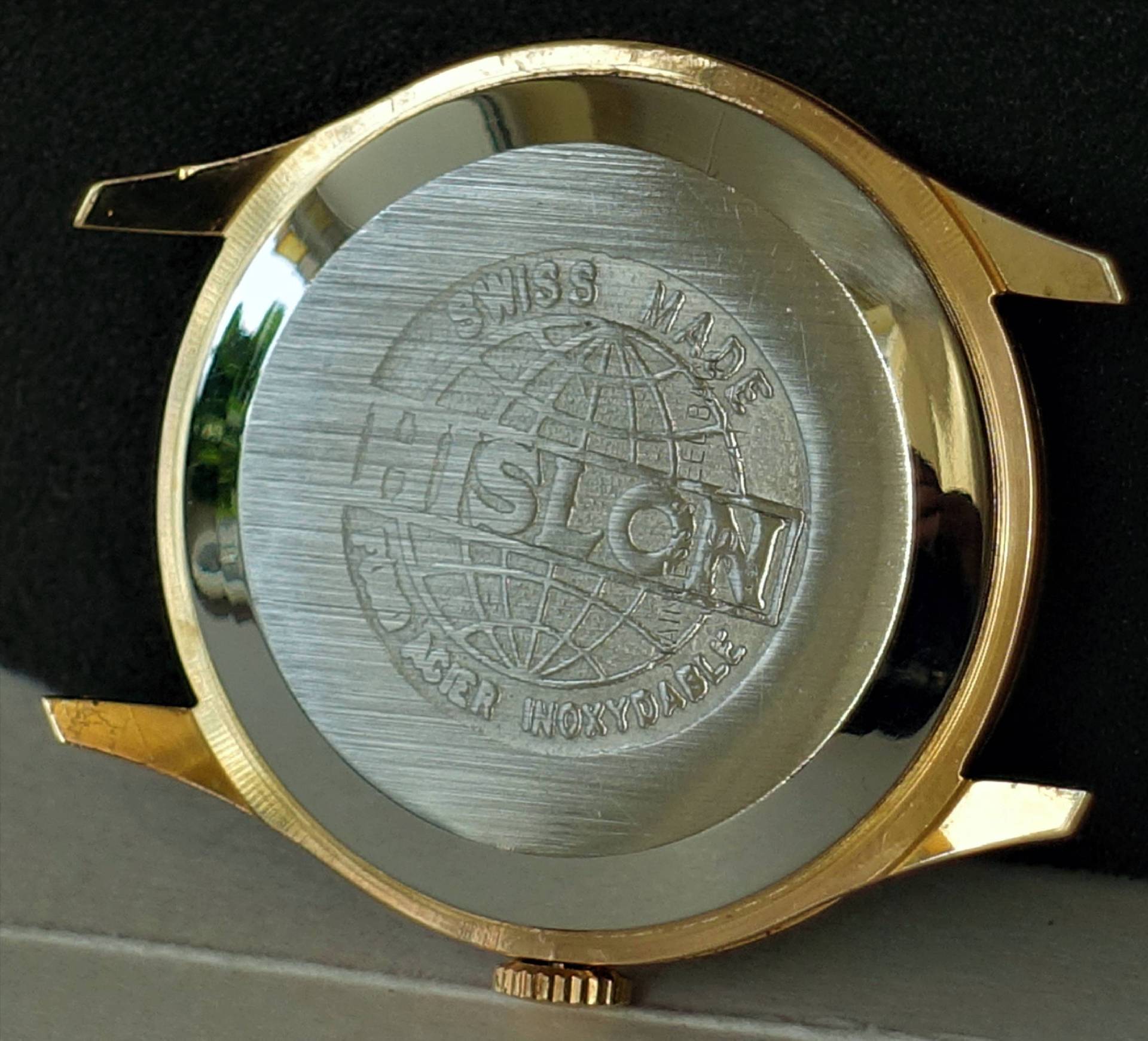 Die Rückseite eines Uhrgehäuses in silberner und goldfarbener Optik mit Gravur des Markennamens und einer Weltkugel.