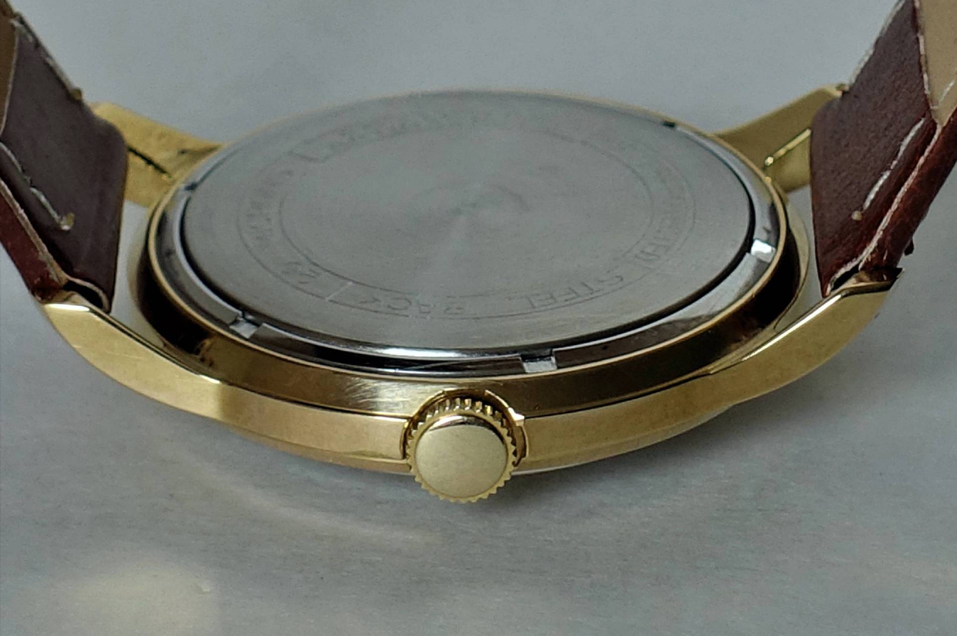 Rückseite einer goldfarbenen Armbanduhr mit braunem Armband und silberfarbenem Gehäuseboden.