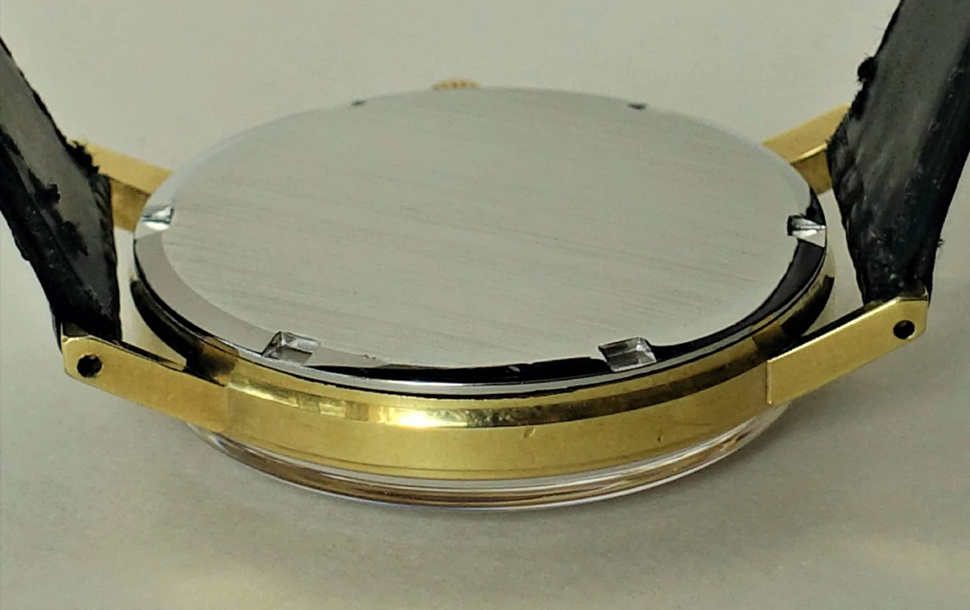 Runder, silberfarbener Uhrenboden an einem goldfarbenen Uhrengehäuse mit schwarzem Armband.