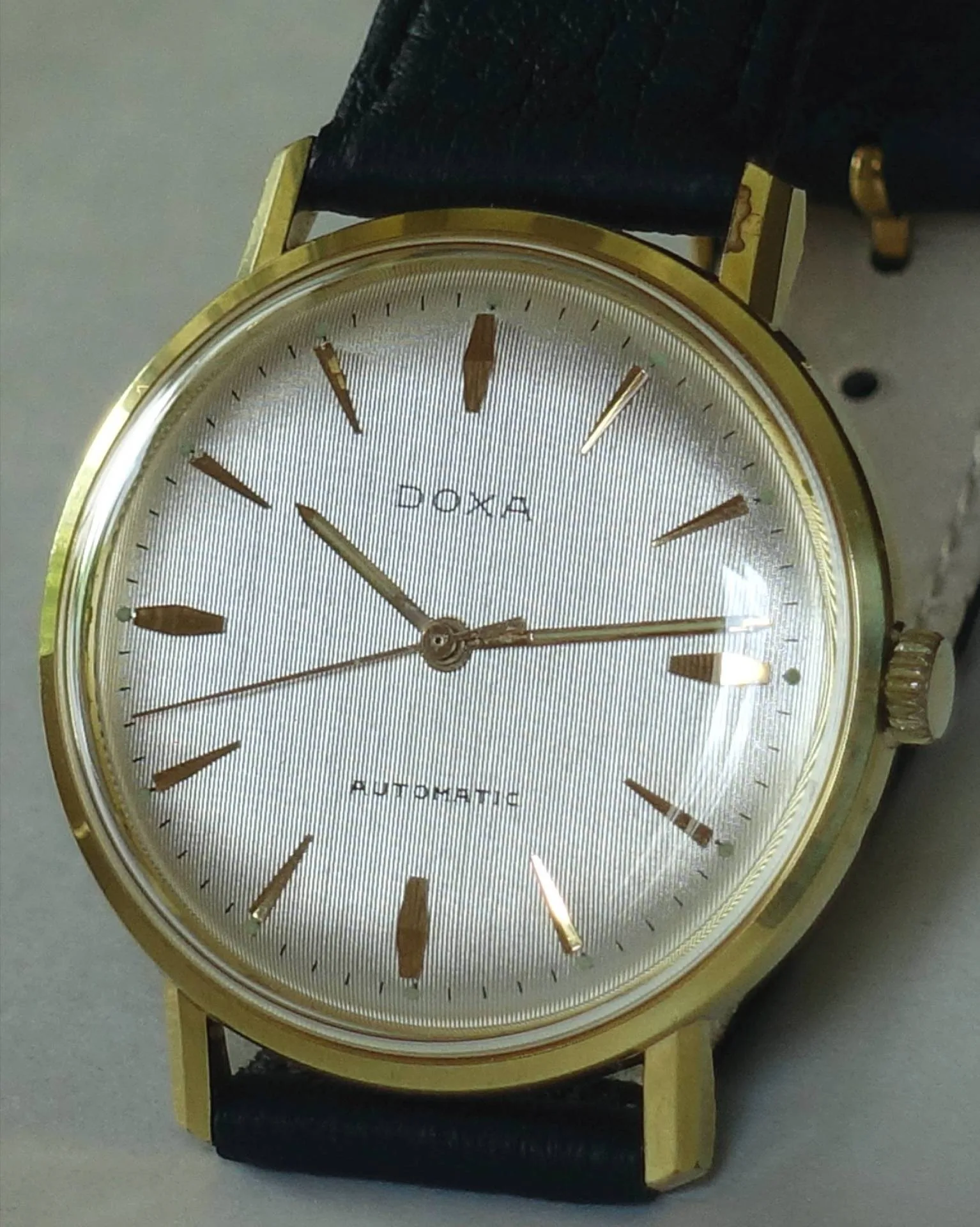 Goldfarbene Doxa Armbanduhr mit hellem Zifferblatt und schwarzem Armband.