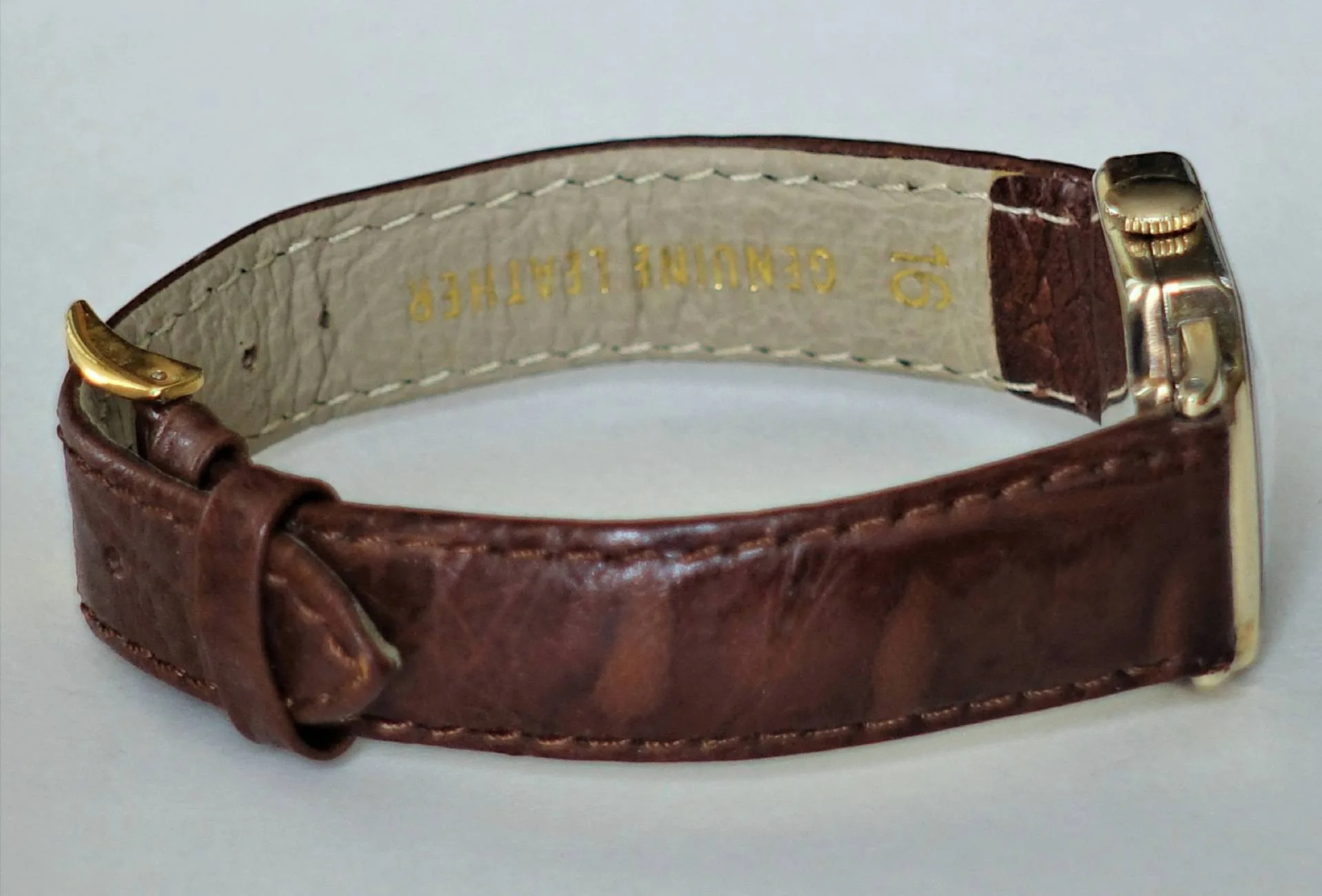 Braunes Lederband, gelb-metallene Schließe Eine Armbanduhr mit braunem Lederarmband und einer gelb-metallfarbenen Schließe liegt seitlich, sodass das Innenfutter mit der Aufschrift "GENUINE LEATHER" sichtbar ist.