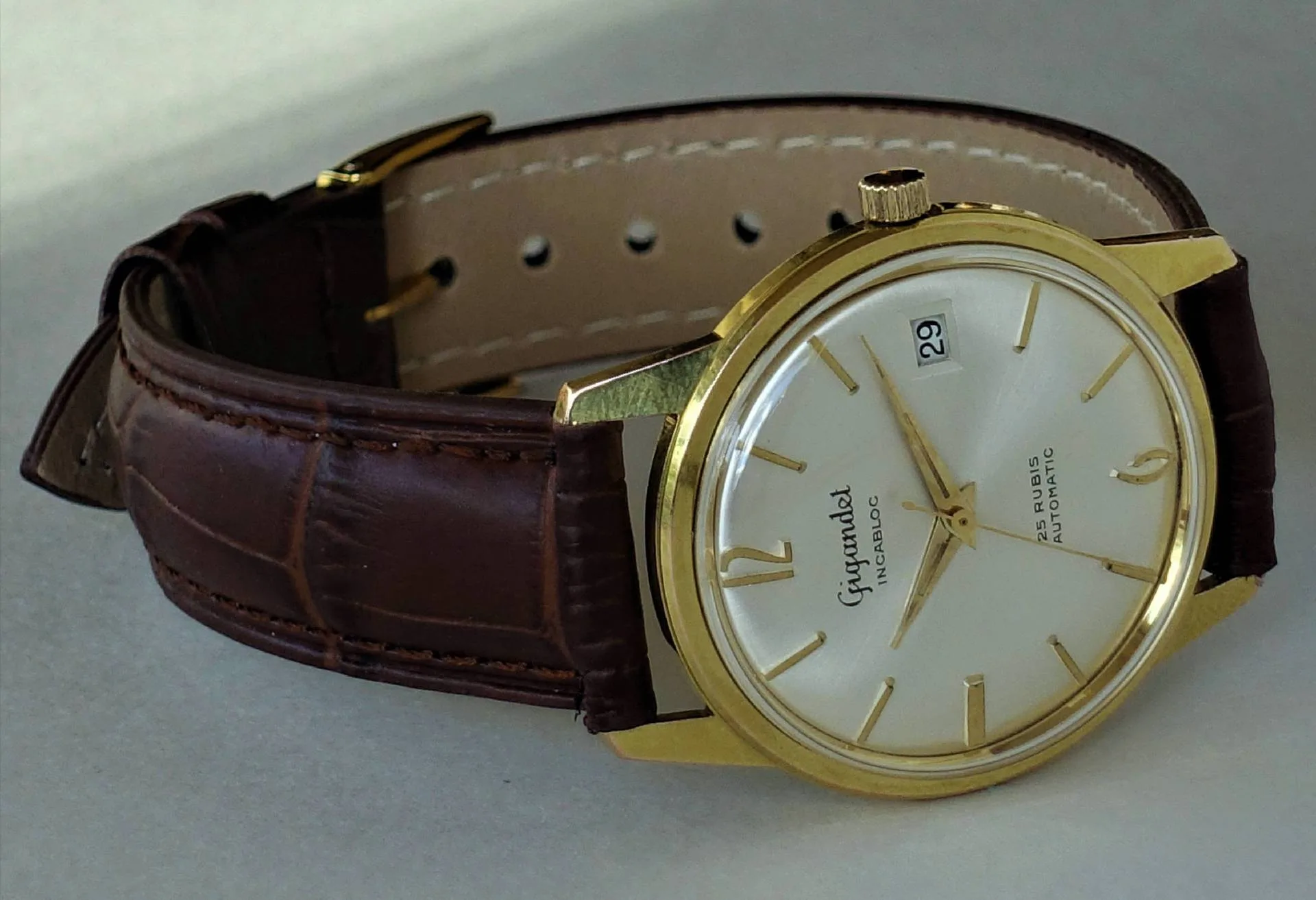 Eine klassische Armbanduhr mit goldfarbenem Gehäuse, hellem Zifferblatt und braunem Lederarmband liegt auf einer hellen Oberfläche.