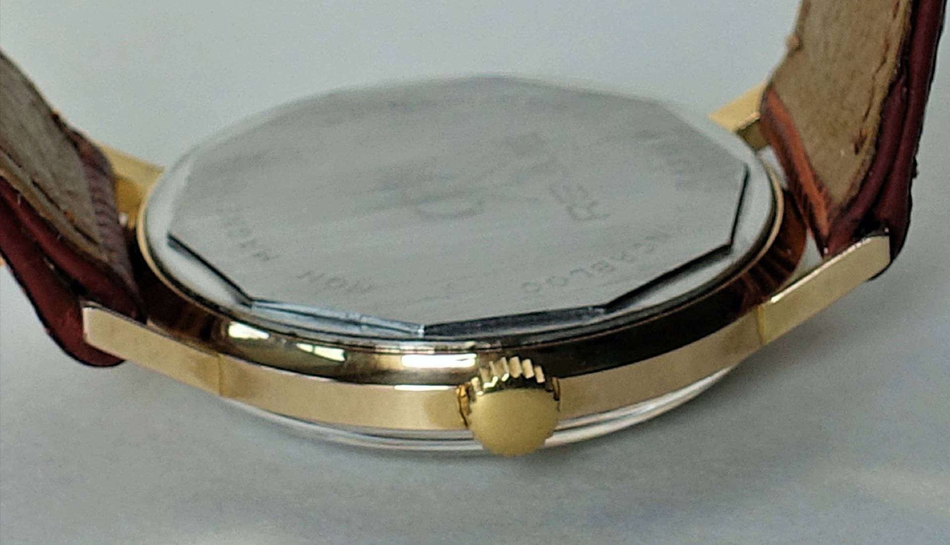 Flache Armbanduhr mit goldfarbenem Gehäuse, braunem Lederarmband und silberfarbener Rückseite.