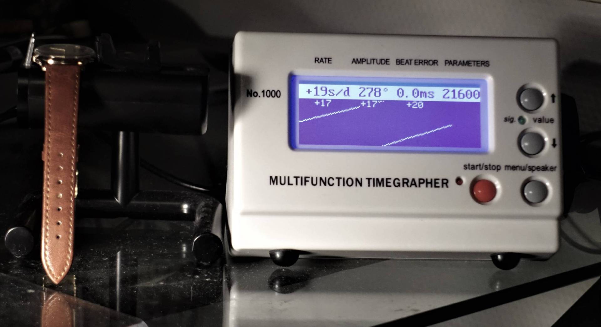 Eine beige Armbanduhr mit braunem Lederarmband liegt auf einem Mikrofonständer neben einem Multifunktions-Timegrapher, dessen Display Messwerte einer mechanischen Uhr anzeigt.