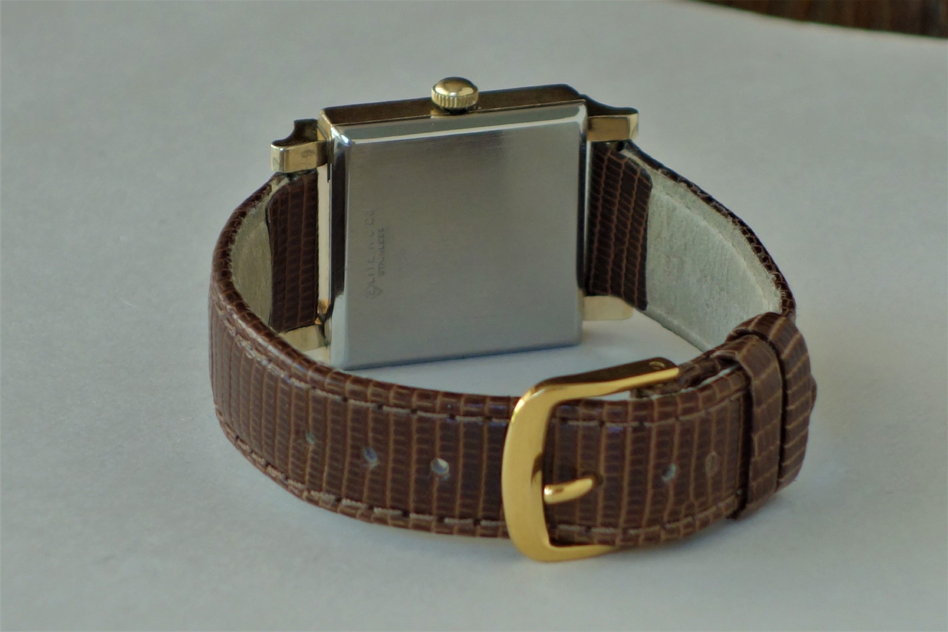Rechteckige Armbanduhr mit braunem Lederarmband und goldfarbener Schließe, Rückseite aus Metall.