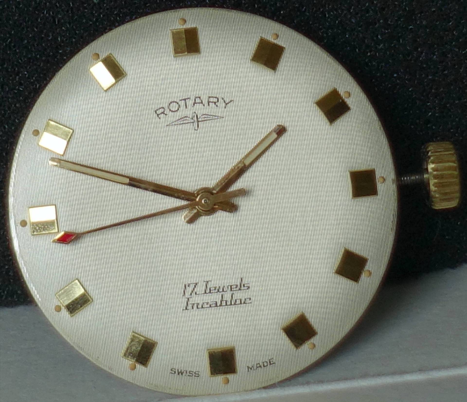 Rundes Zifferblatt einer Rotary-Uhr mit goldfarbenen Zeigern und Indizes auf hellem Hintergrund.