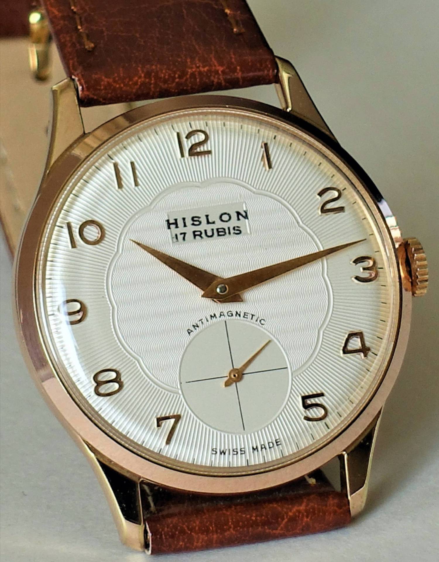 Eine Armbanduhr mit weißem Zifferblatt, goldfarbenem Gehäuse und braunem Lederarmband, zeigt die Marke "HISLON" und eine kleine Sekunde bei sechs Uhr.