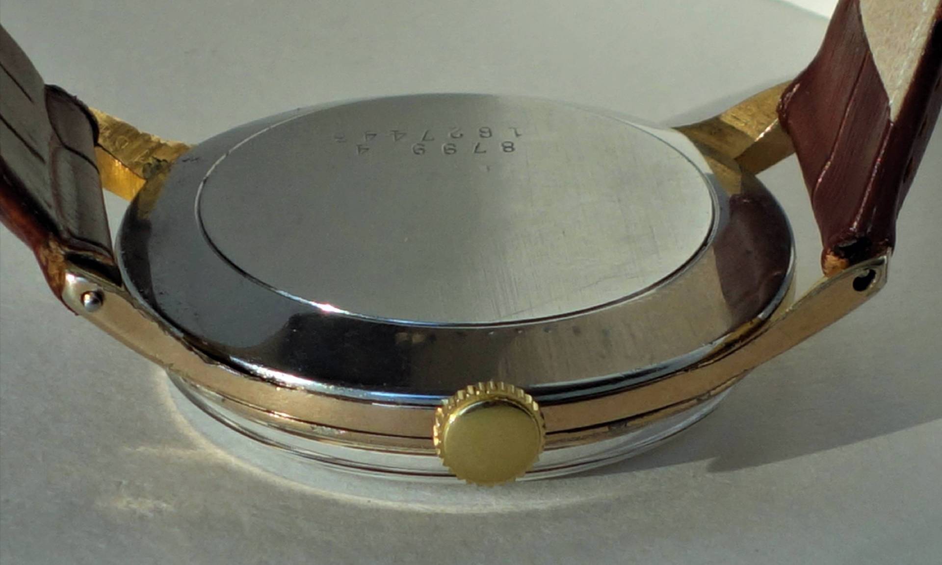 Silberfarbener Uhrenboden mit braunem Armband und goldfarbener Krone.