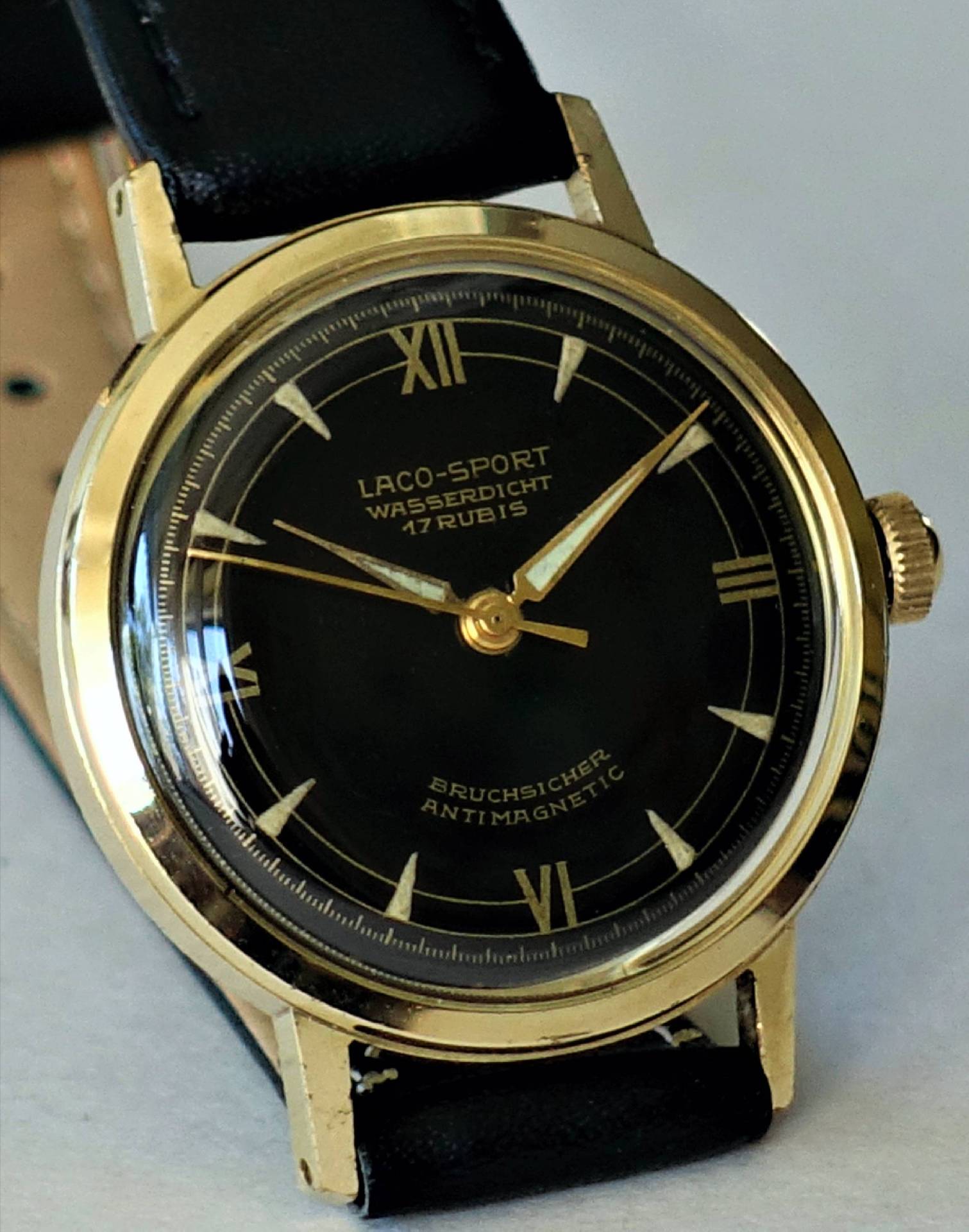 Schwarze Armbanduhr mit goldfarbenem Gehäuse und schwarzem Lederarmband, Zifferblatt mit römischen Zahlen und goldener Beschriftung.