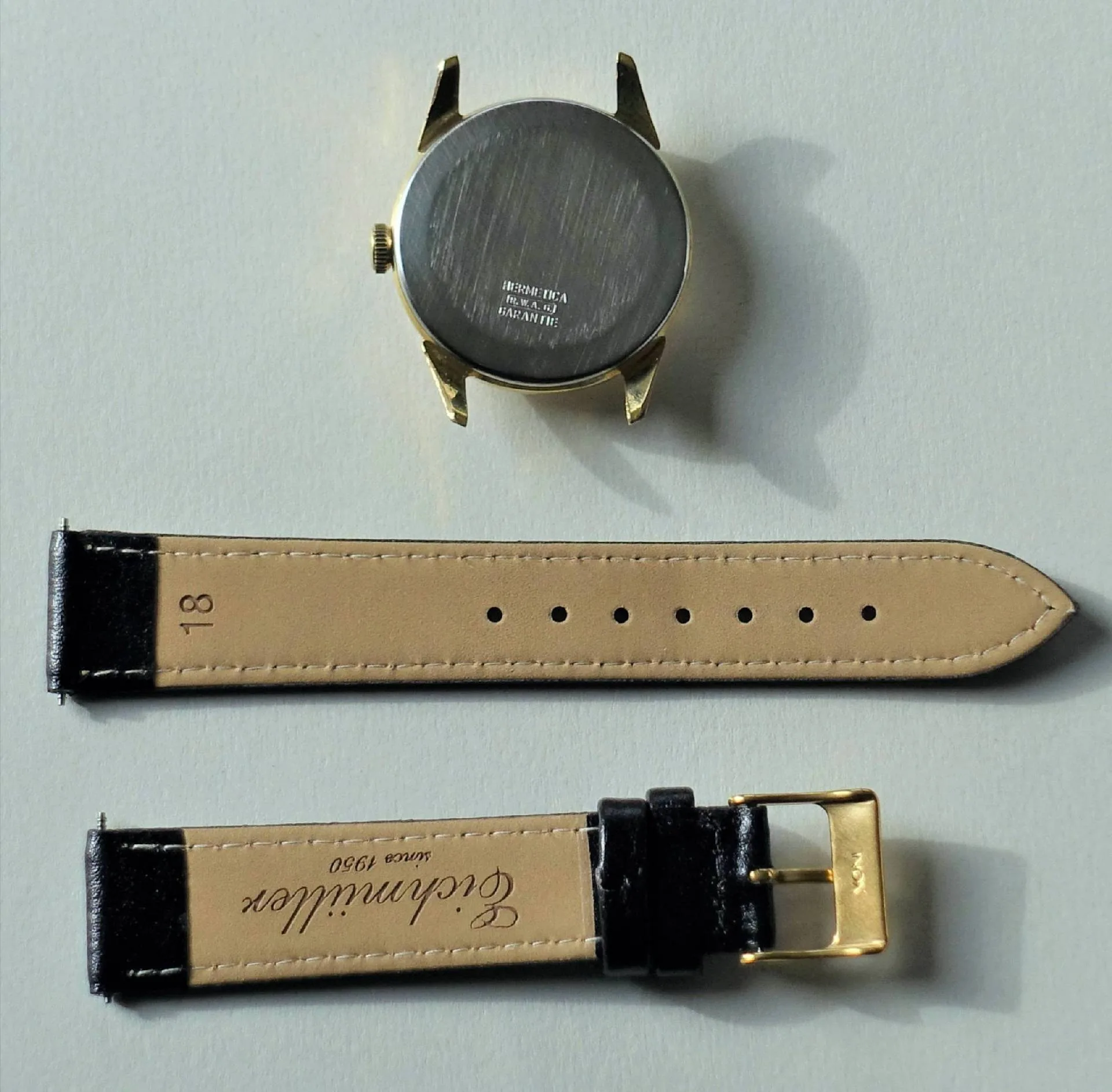 Goldfarbenes Uhrgehäuse mit schwarzem Lederarmband und goldfarbener Schließe, separat abgebildet.