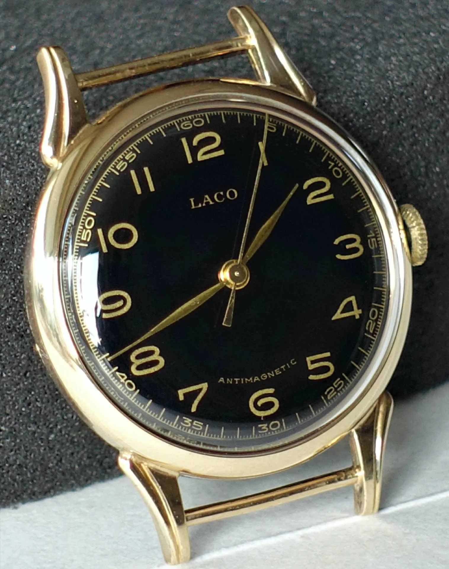 Eine goldfarbene Armbanduhr mit schwarzem Zifferblatt und goldenen Zahlen und Zeigern, ohne Armband, auf dunklem Hintergrund.