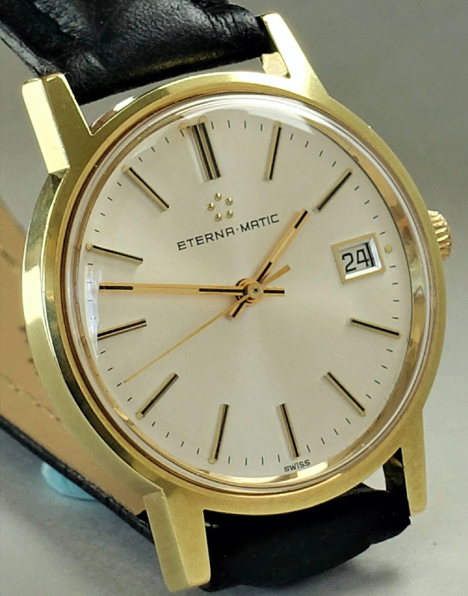 Goldfarbene Armbanduhr mit hellem Zifferblatt, schwarzem Lederarmband und Datumsanzeige.