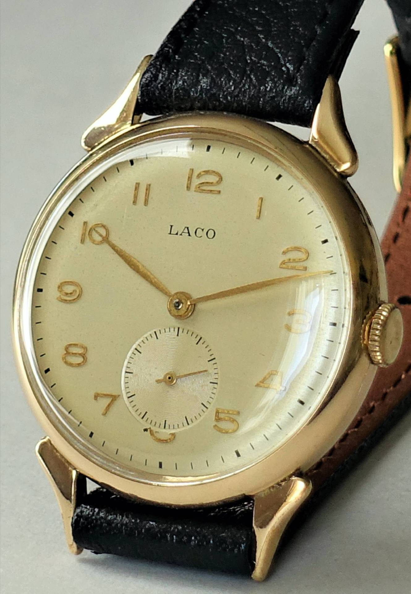 Goldfarbene Armbanduhr mit hellem Zifferblatt, schwarzen Zahlen und schwarzem Lederarmband.