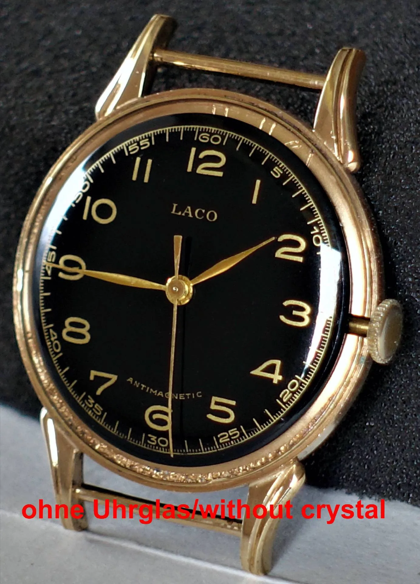 Eine Armbanduhr mit schwarzem Zifferblatt, goldfarbenen Zeigern und Gehäuse, ohne Uhrglas, liegt auf einer dunklen Unterlage.