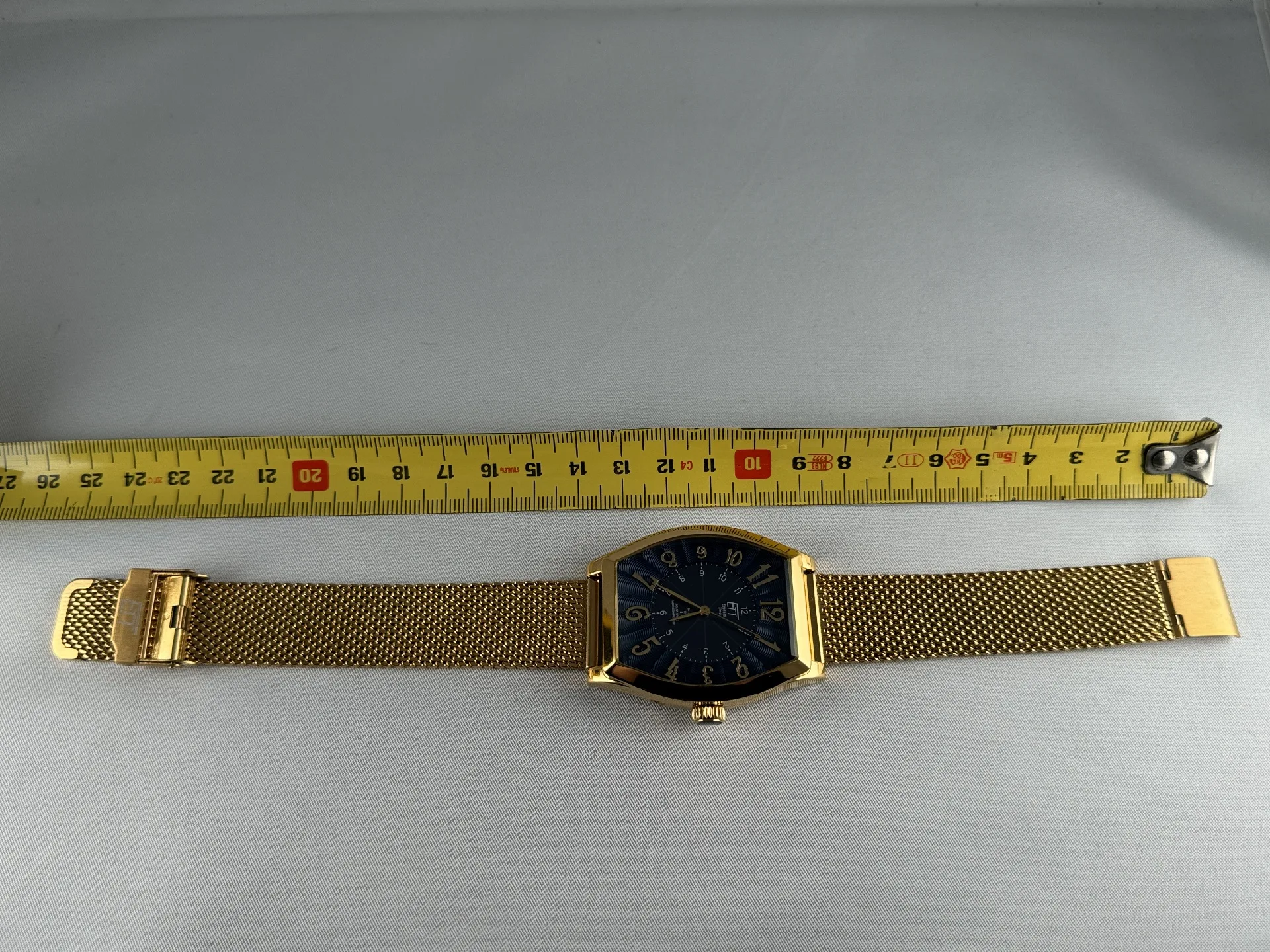 Goldene Armbanduhr Milanaiseband Goldfarbene Armbanduhr mit schwarzem Zifferblatt und Milanaise-Edelstahlarmband, daneben ein Maßband zur Längenmessung.