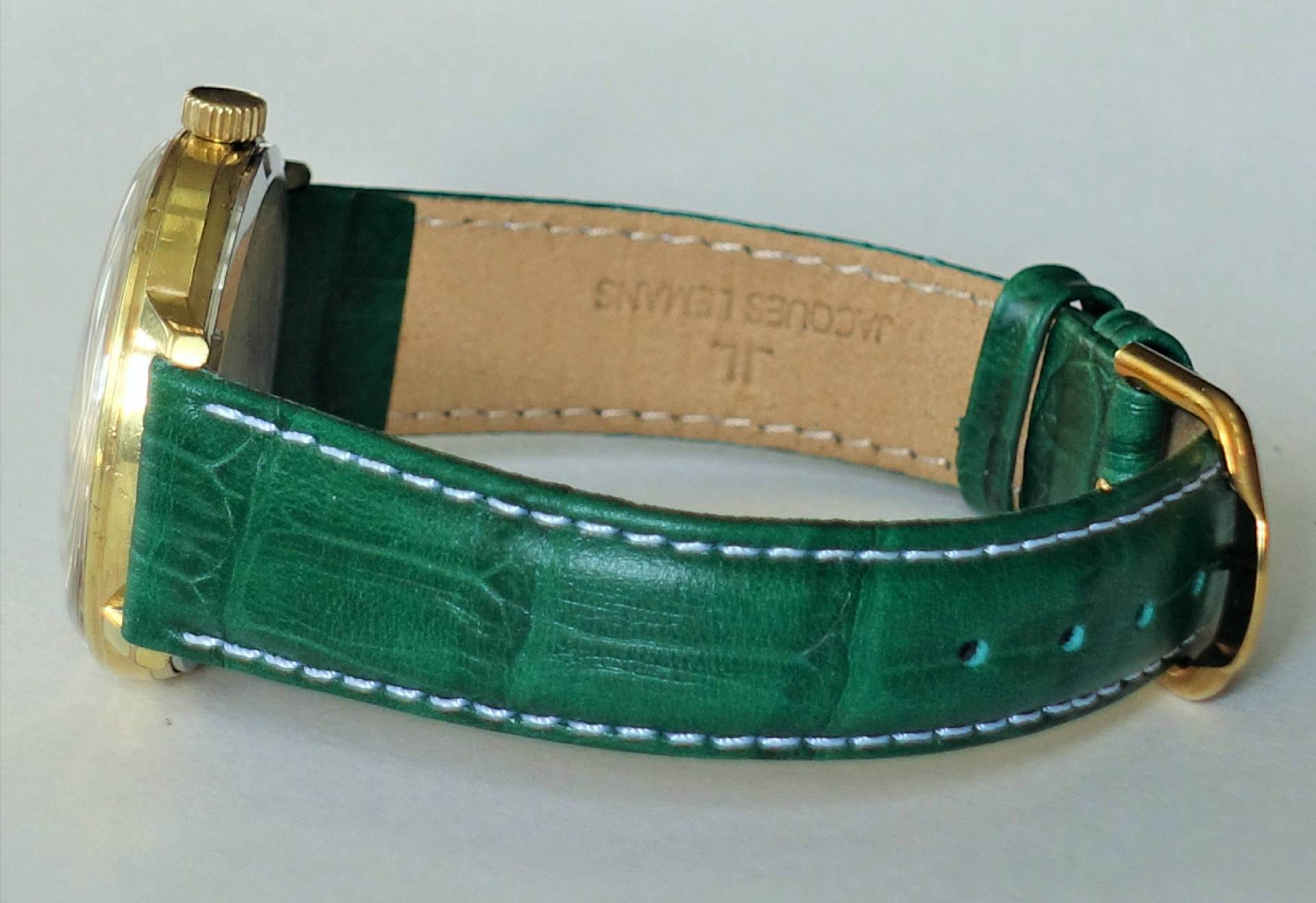Eine Armbanduhr mit grünem Lederarmband, weißer Naht und goldfarbener Schließe liegt seitlich auf einer hellen Oberfläche.