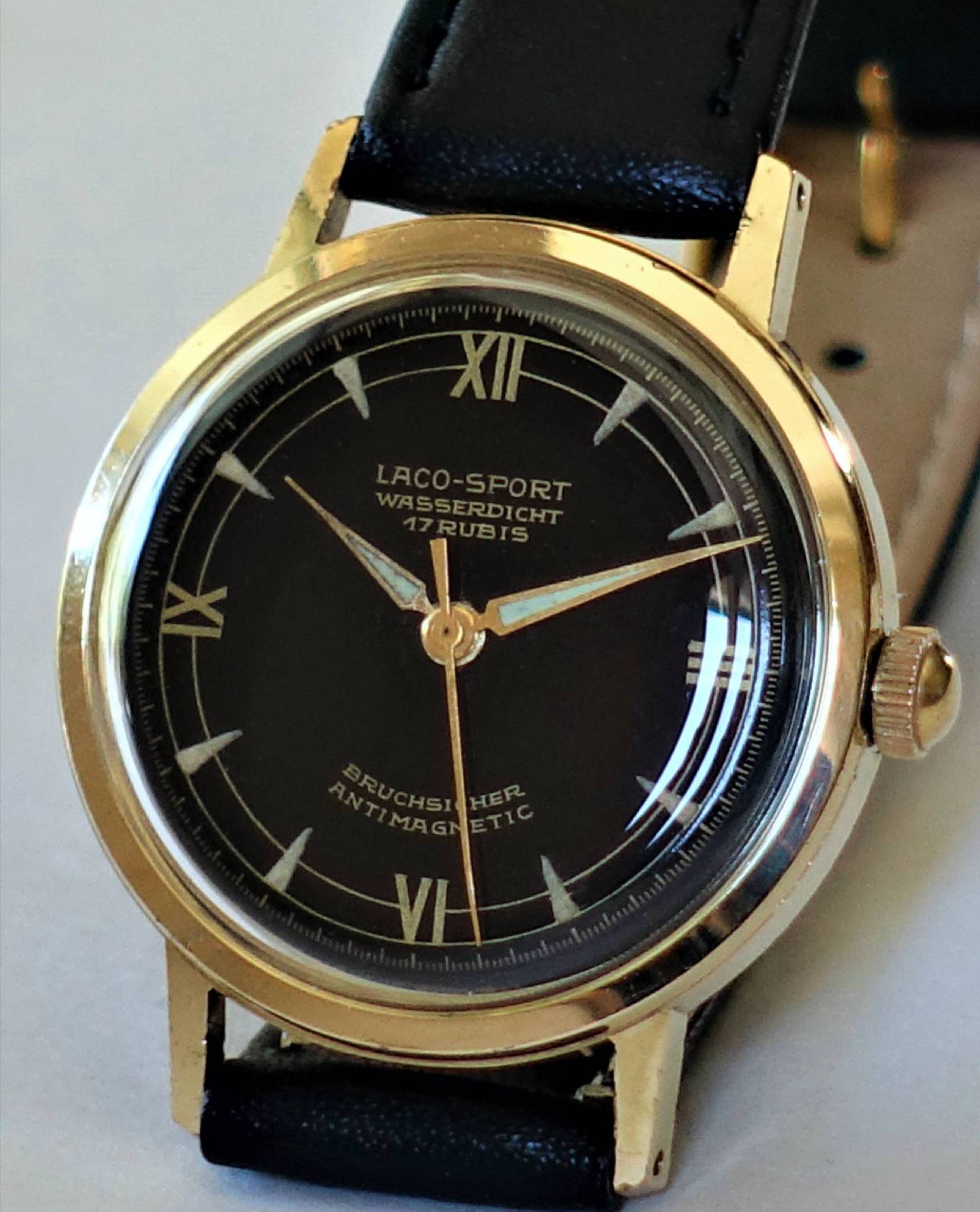 Schwarze Armbanduhr mit goldfarbenem Gehäuse und schwarzem Lederarmband, Zifferblatt mit römischen Zahlen.