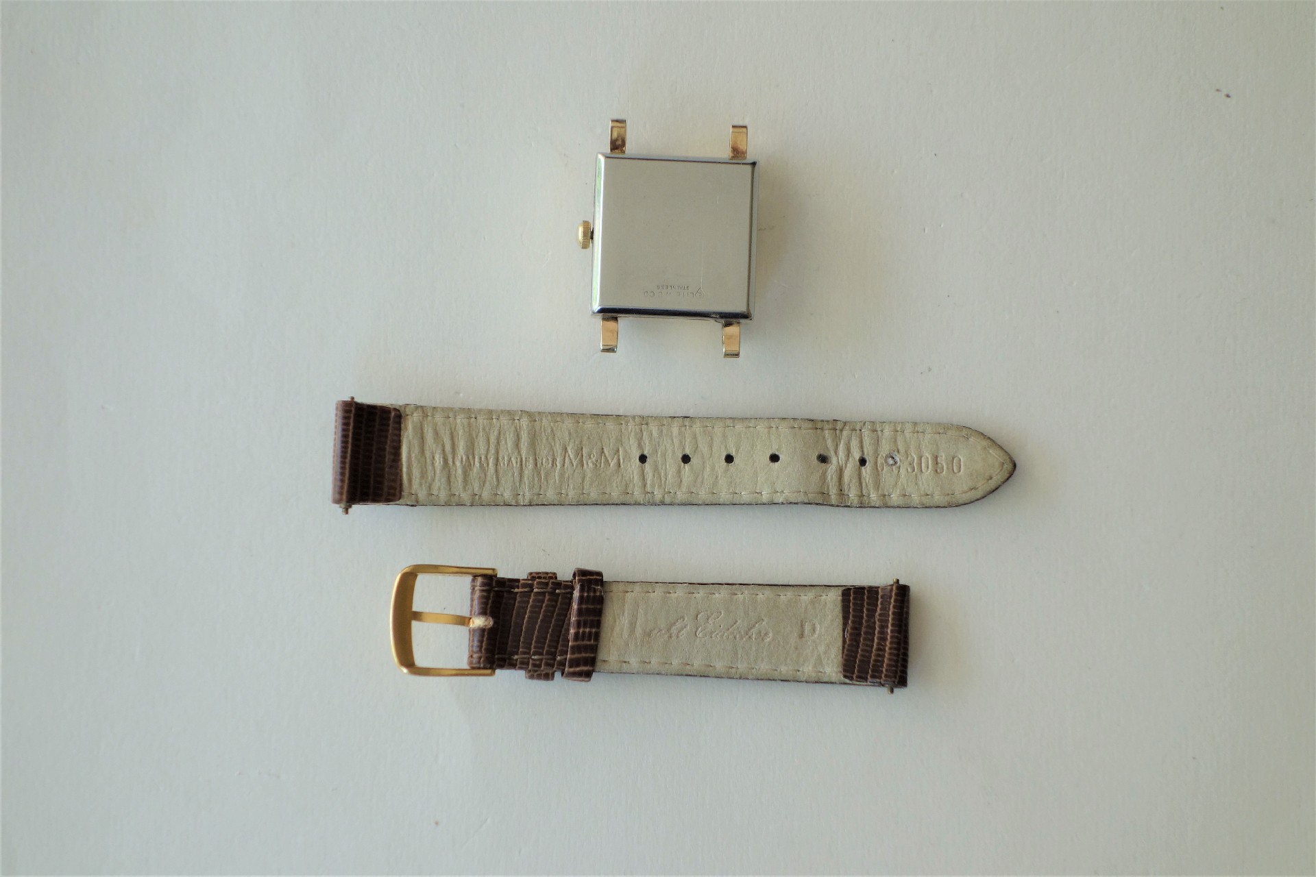 Quadratisches Metall-Uhrgehäuse mit zwei braunen Lederarmbandteilen und goldfarbener Schließe.