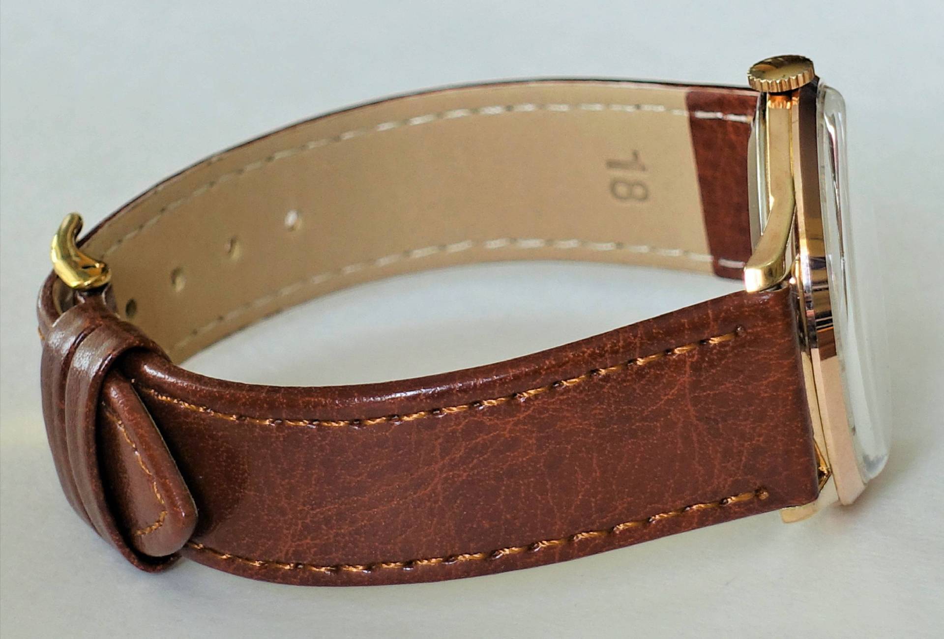 Eine Armbanduhr mit braunem Lederarmband und goldfarbenem Gehäuse liegt seitlich auf einer hellen Oberfläche.