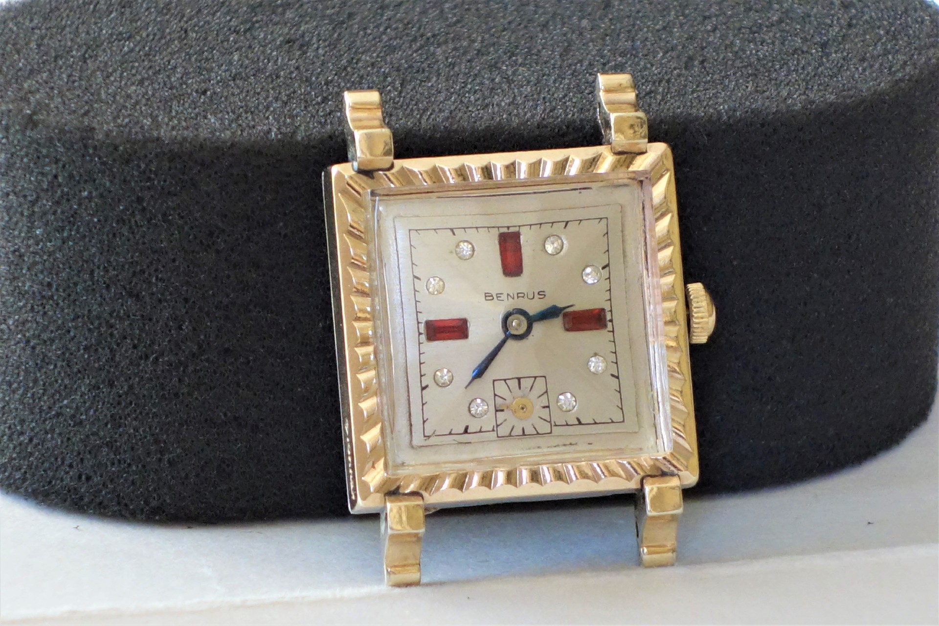 Rechteckige Armbanduhr mit hellem Zifferblatt, goldfarbenem Gehäuse und roten sowie silbernen Indizes.