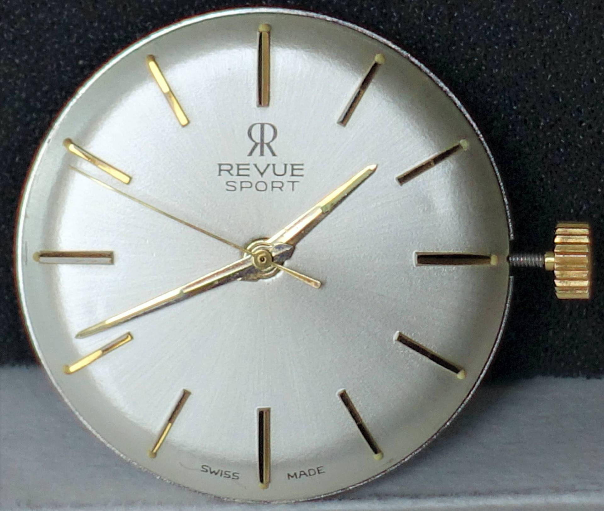 Rundes silberfarbenes Uhrwerk mit goldfarbenen Zeigern und Indizes, sichtbarer Krone, Aufschrift "Revue Sport".