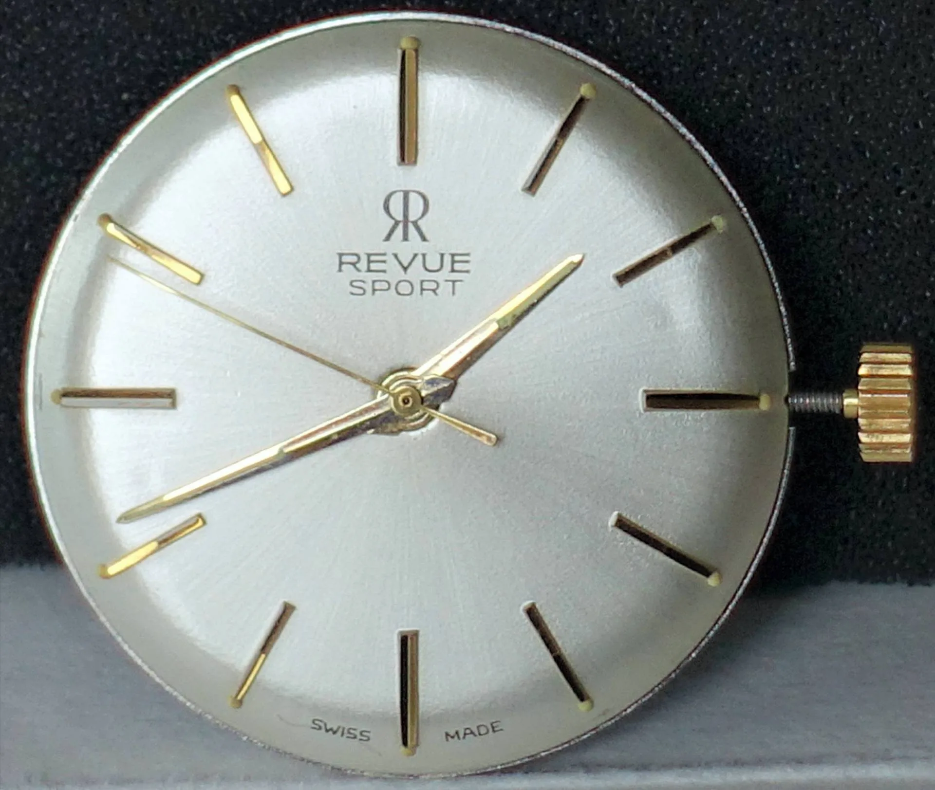 Rundes silberfarbenes Uhrwerk mit goldfarbenen Zeigern und Indizes, sichtbarer Krone, Aufschrift "Revue Sport".