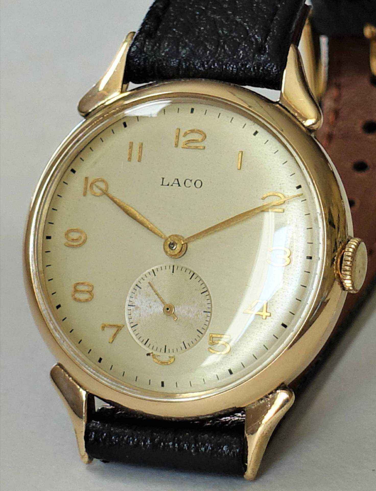 Goldfarbene Armbanduhr mit hellem Zifferblatt, schwarzen Zahlen und schwarzem Lederarmband.