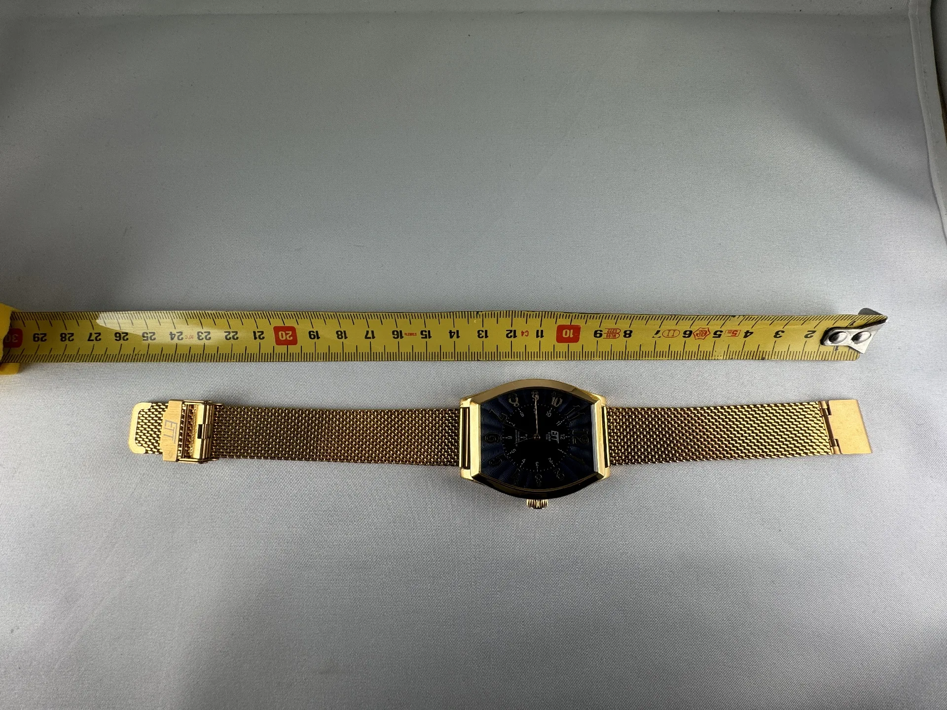 Goldfarbene Armbanduhr mit schwarzem Zifferblatt und Milanaise-Edelstahlarmband, daneben ein Maßband zur Längenmessung.