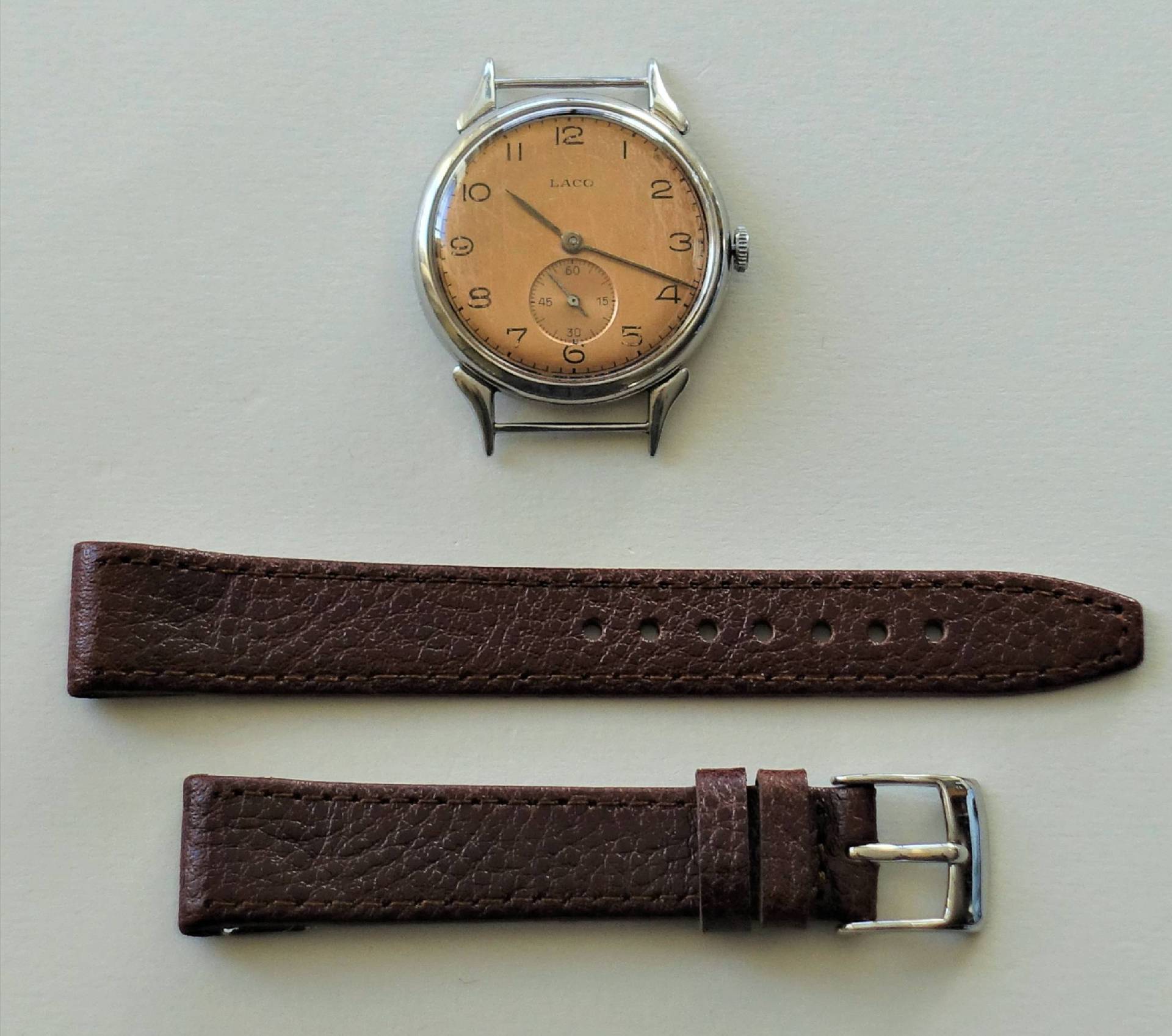 Silberfarbene Armbanduhr mit beigem Zifferblatt und separat liegendem braunem Armband aus Leder.