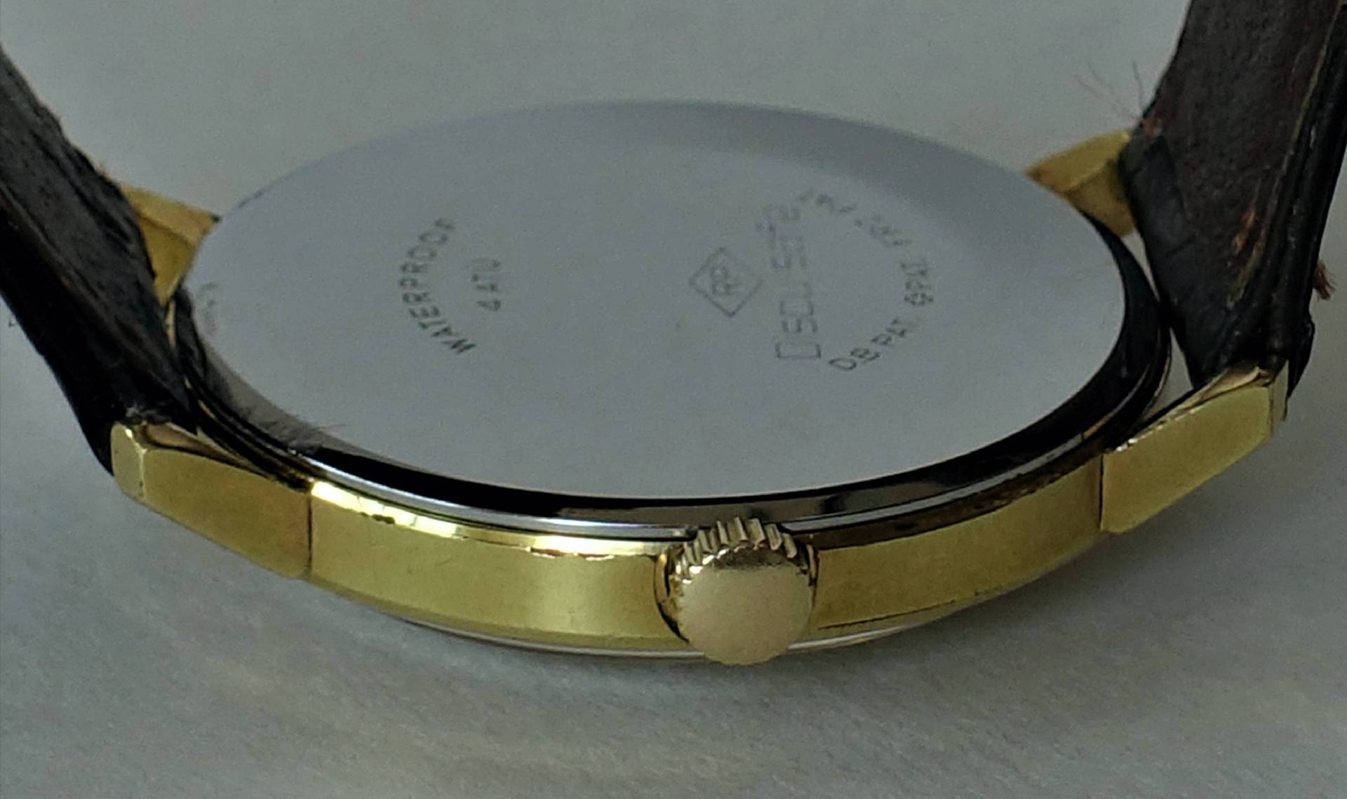 Runder, goldfarbener Uhrenboden mit seitlicher Krone und schwarzem Lederarmband.