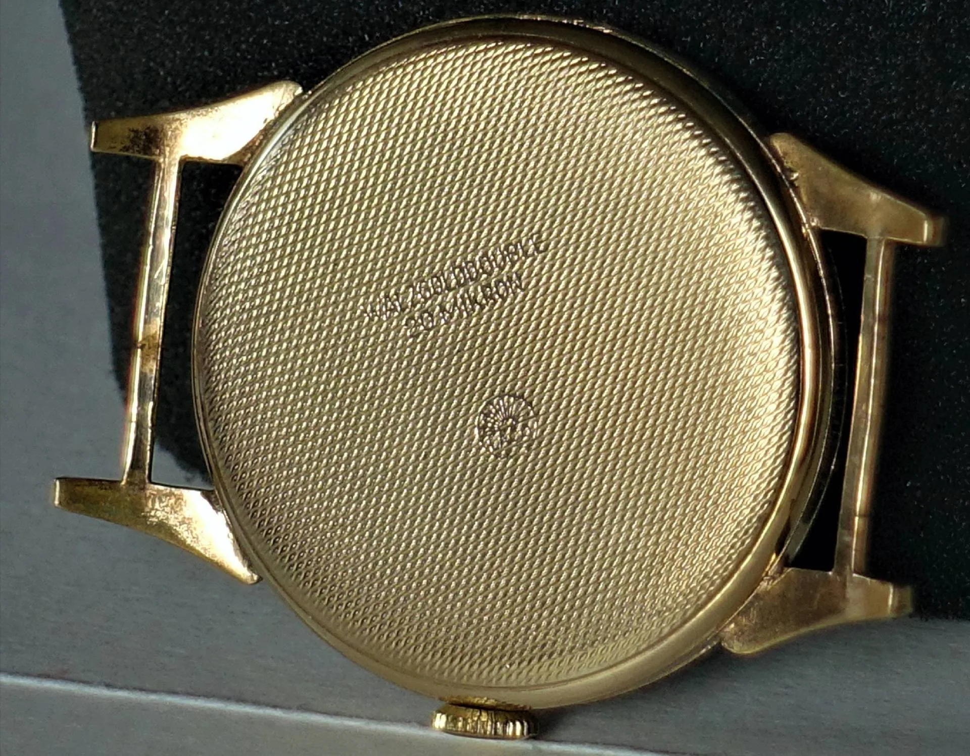 Rückseite eines gelb-metallfarbenen, fein strukturierten Uhrgehäuses ohne Armband, mit Gravur in der Mitte.